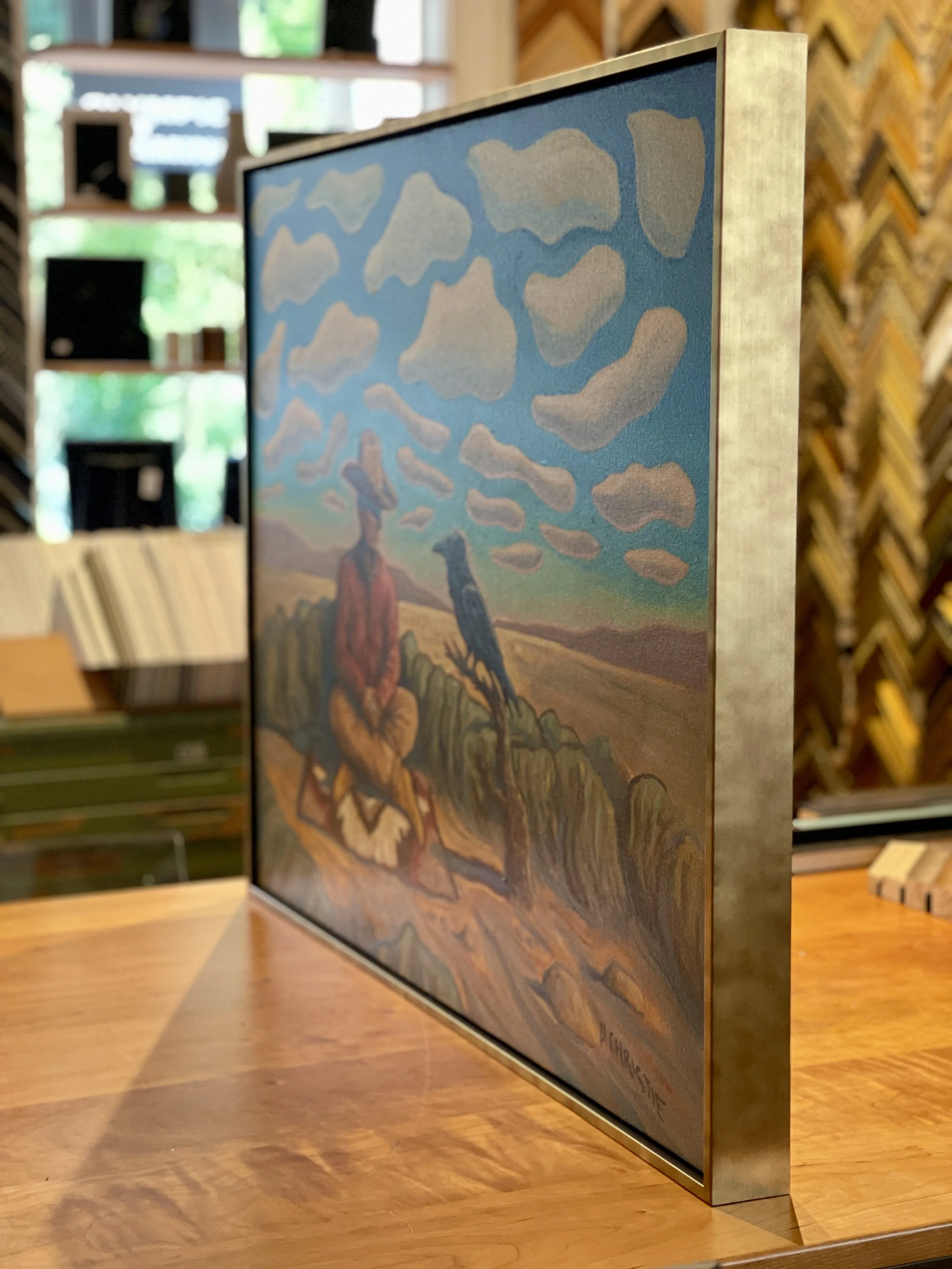 A framed painting of a person sitting outdoors with two birds on a branch, under a sky filled with clouds, displayed on a wooden table in an art store.