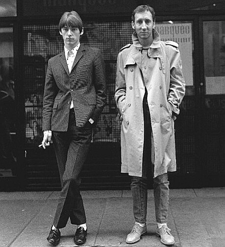 2 men standing on the street, the left in a suit posing with his legs crossed. The one on th eright in a trench coat with his hands in his pockets