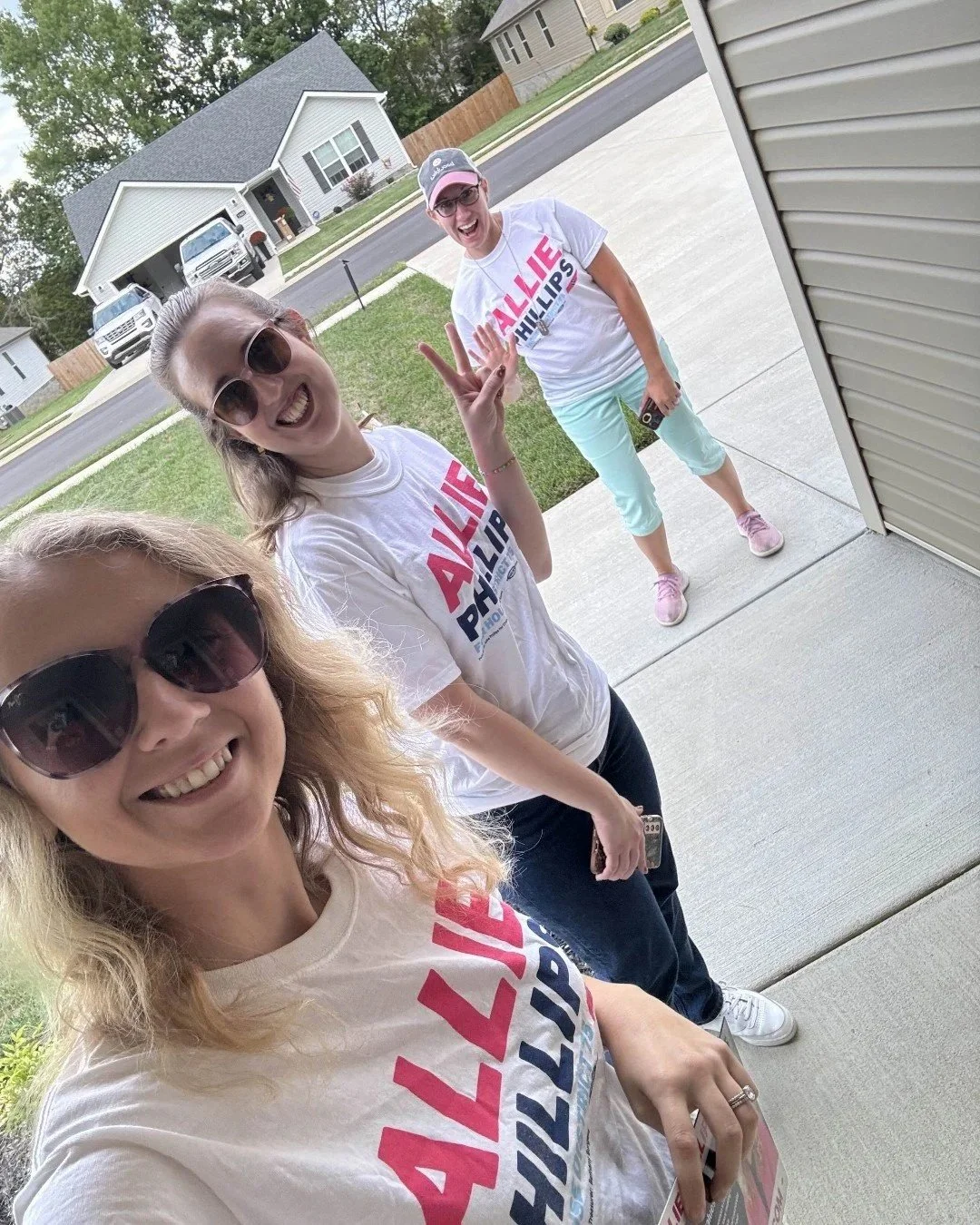 Three women outdoors near a house, taking a selfie while wearing white t-shirts with red and blue text, smiling, and making peace signs.