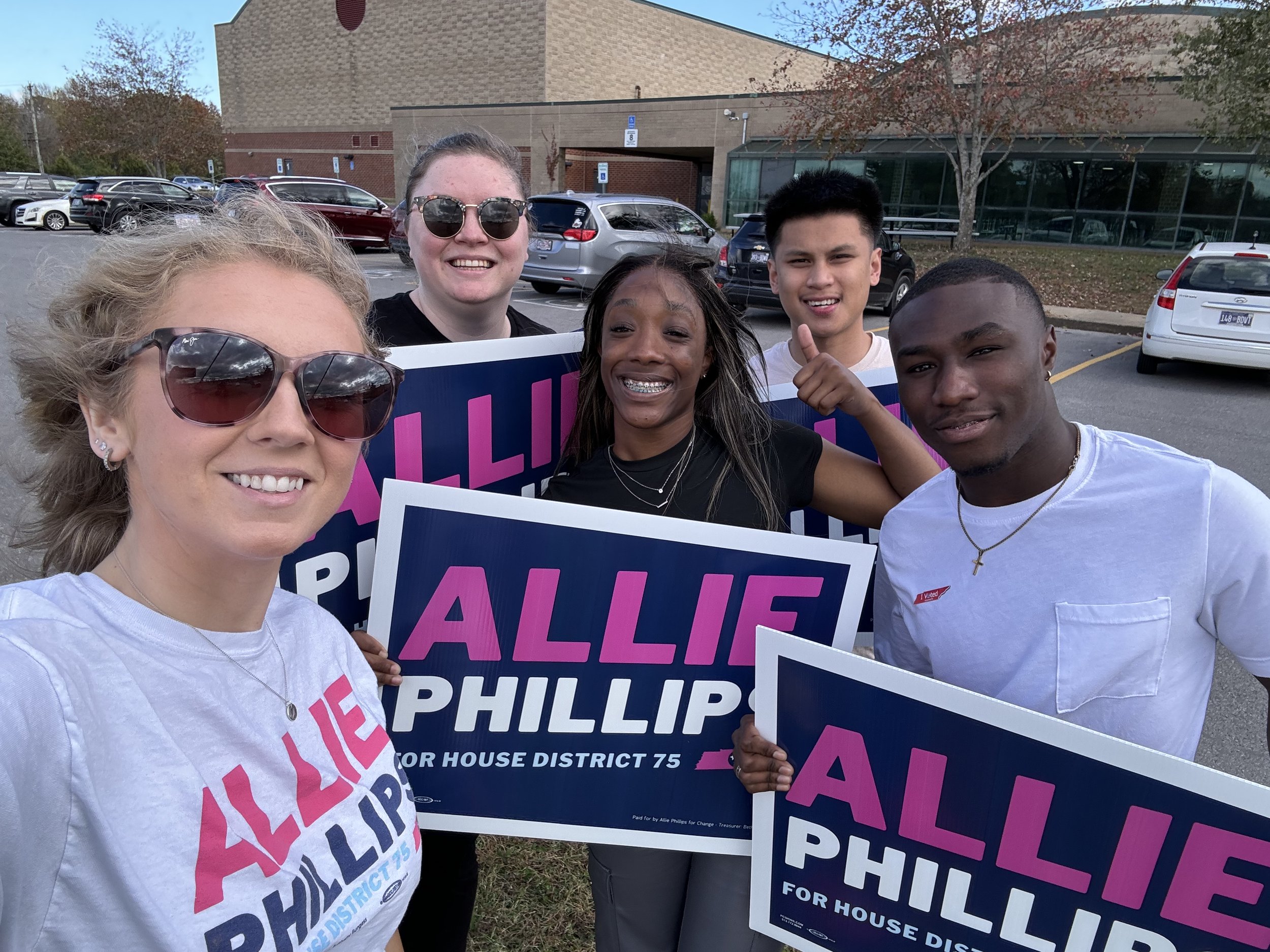 Group of five people smiling and holding campaign signs for Allie Phillips, candidate for House District 75, outdoors in a parking lot with cars and a building in the background.