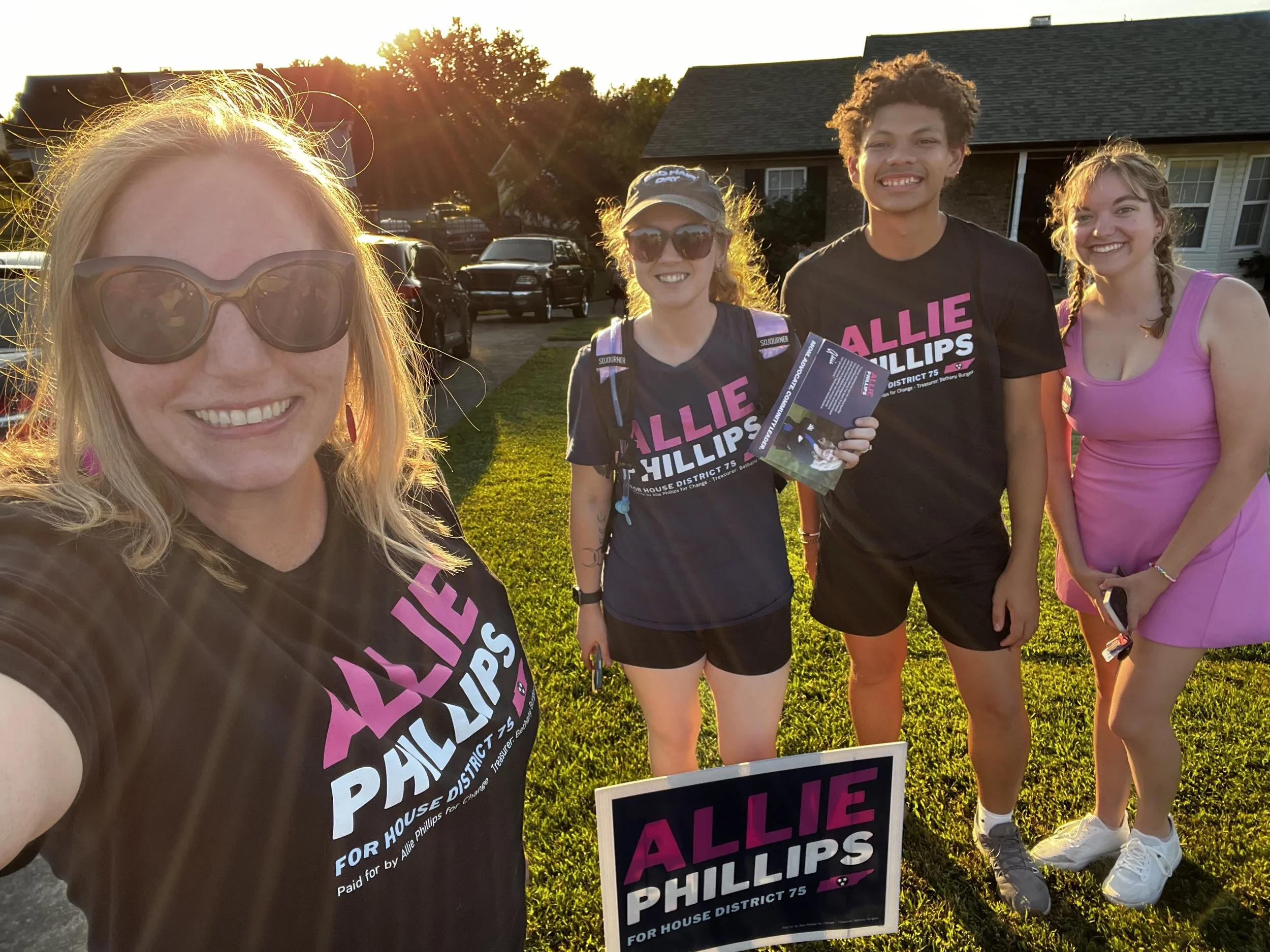 Four people standing outside on a grassy lawn during sunset, wearing black t-shirts with pink and white text supporting Allie Phillips for House District 75, smiling and posing for a photo.