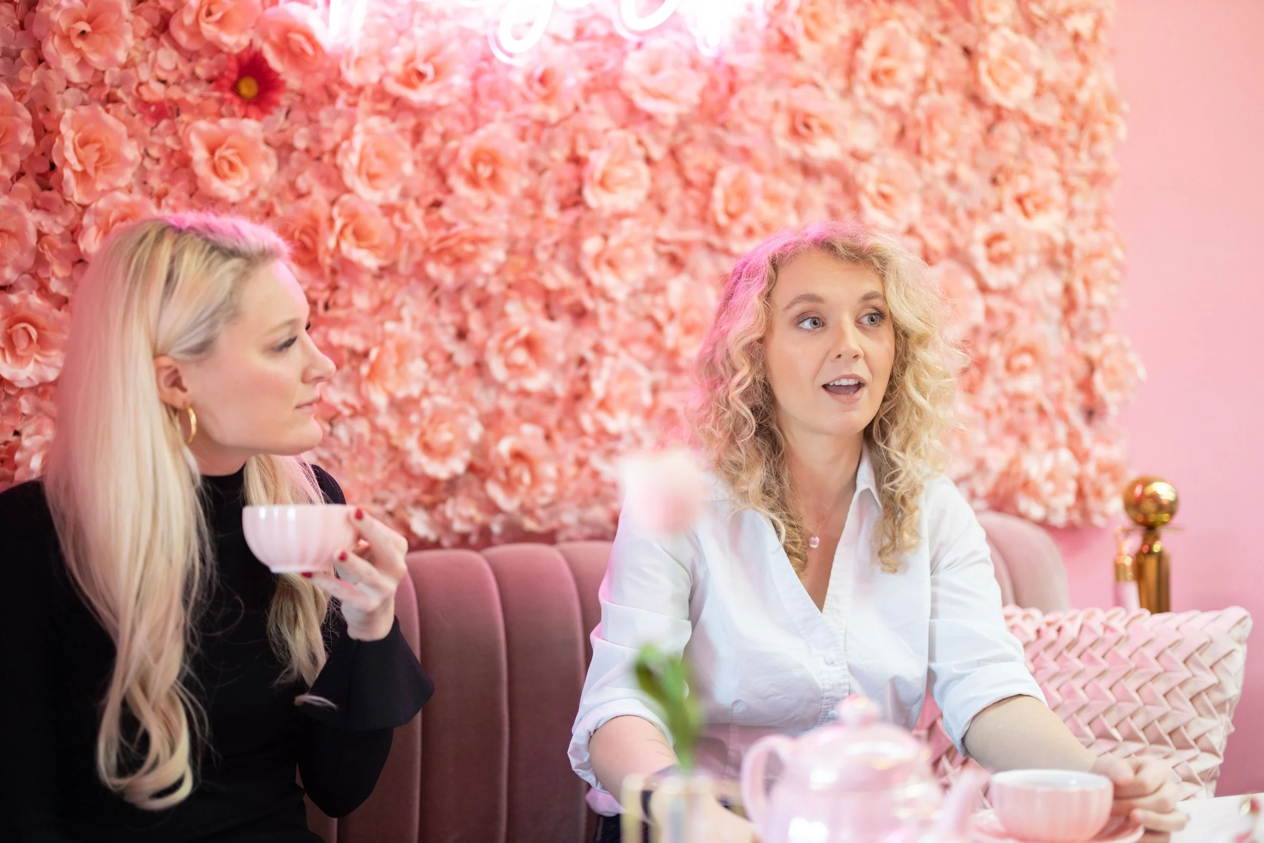 Two women sit at a table in front of a pink floral wall. One woman with blonde wavy hair, wearing a white shirt, looks surprised or engaged. The other woman with long blonde hair, wearing a black top, holds a teacup and looks to the side. The table has pink teapots and cups, and the ambiance suggests a tea party or brunch in a stylish, pink-themed setting.