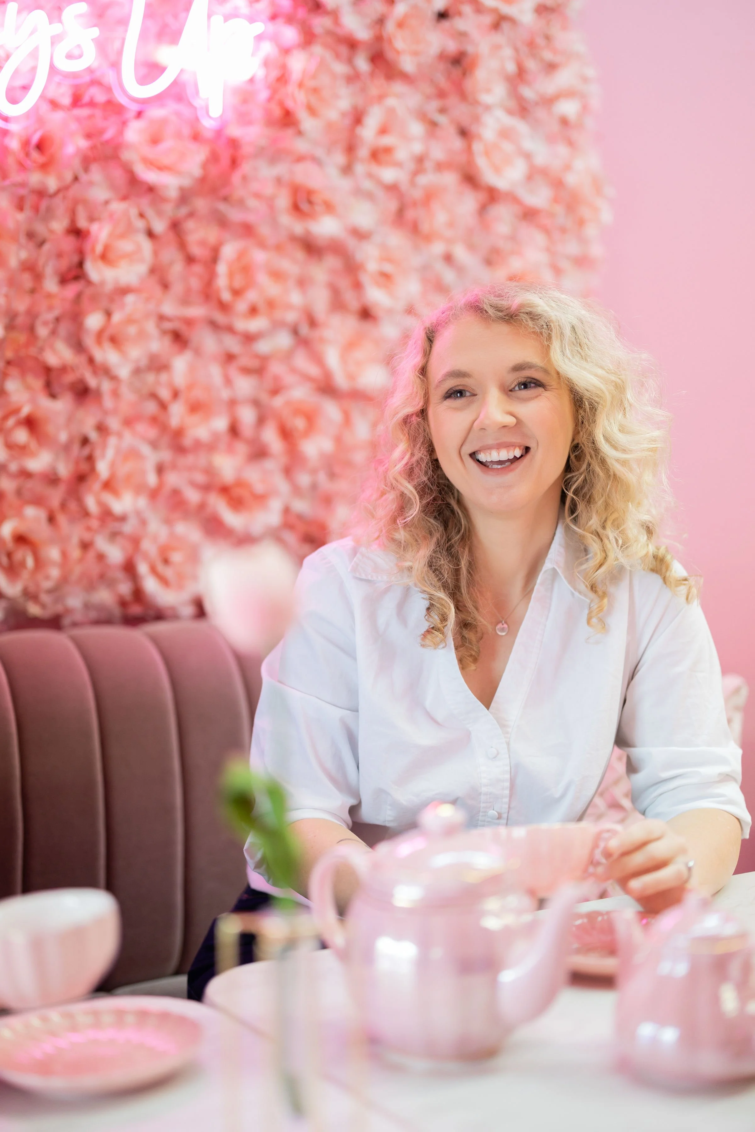 A woman with curly blonde hair smiling, seated at a pink-themed tea or dessert shop with pink teapot and cups, pink flower wall background, and pink neon sign.