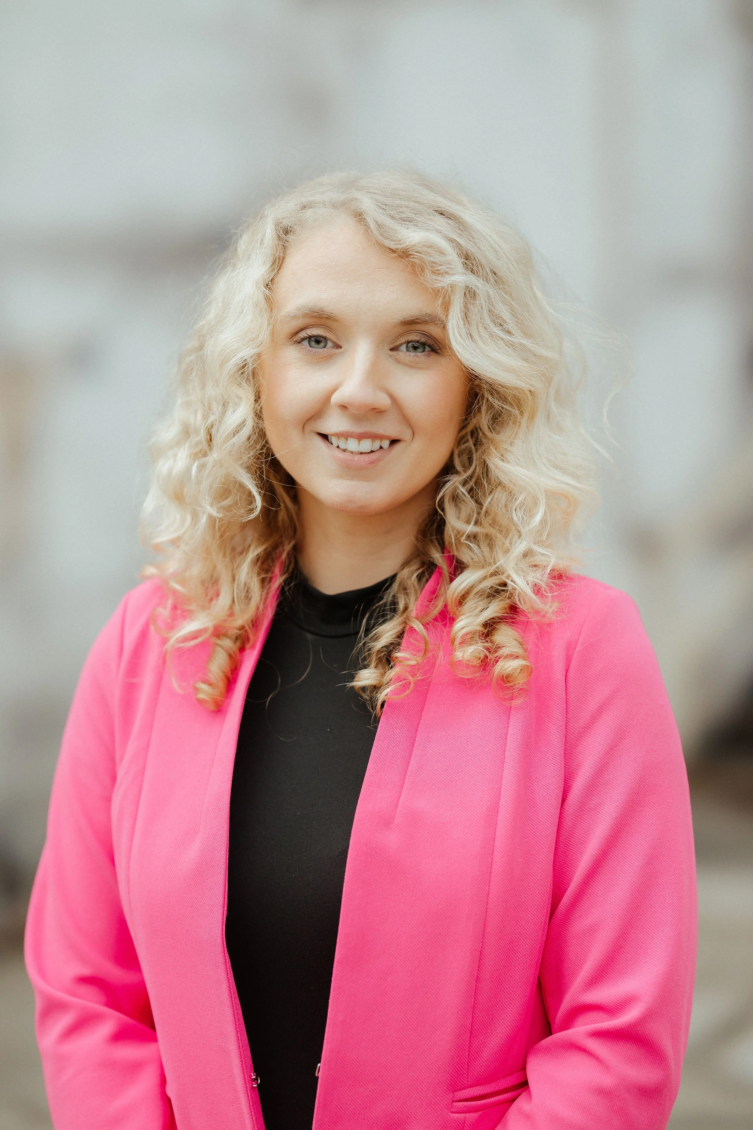 A young woman with blonde curly hair, smiling, wearing a black top and a pink blazer, standing outdoors with a blurred urban background.