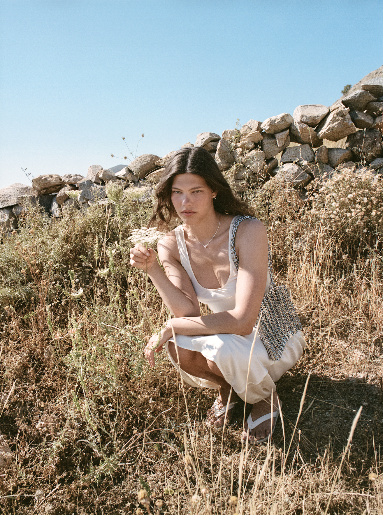 A young woman with long dark hair squats in dry grassy field near a stone wall under clear blue sky, wearing a white dress, colorful sandals, and carrying a patterned tote bag.