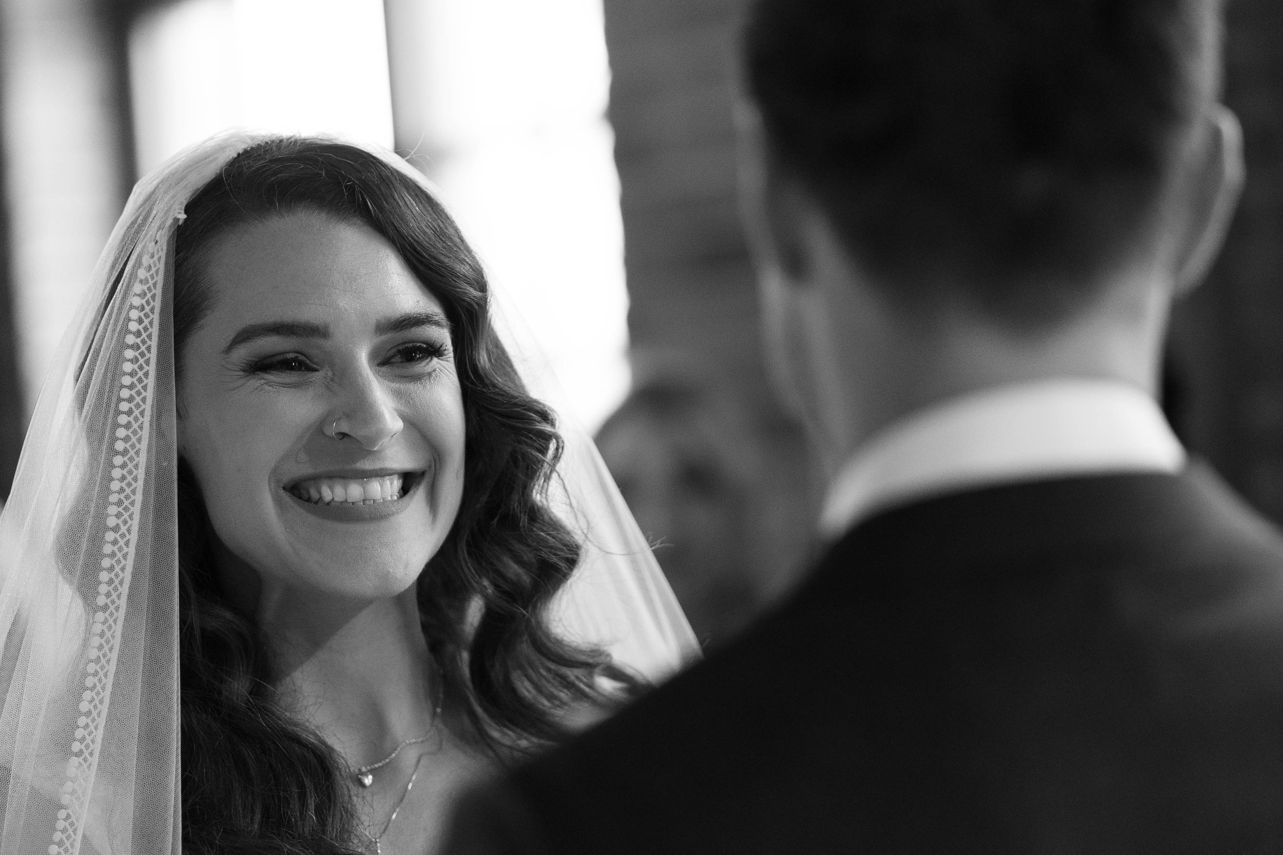 A smiling bride with dark wavy hair, wearing a veil and necklace, looks at her groom during their wedding ceremony.