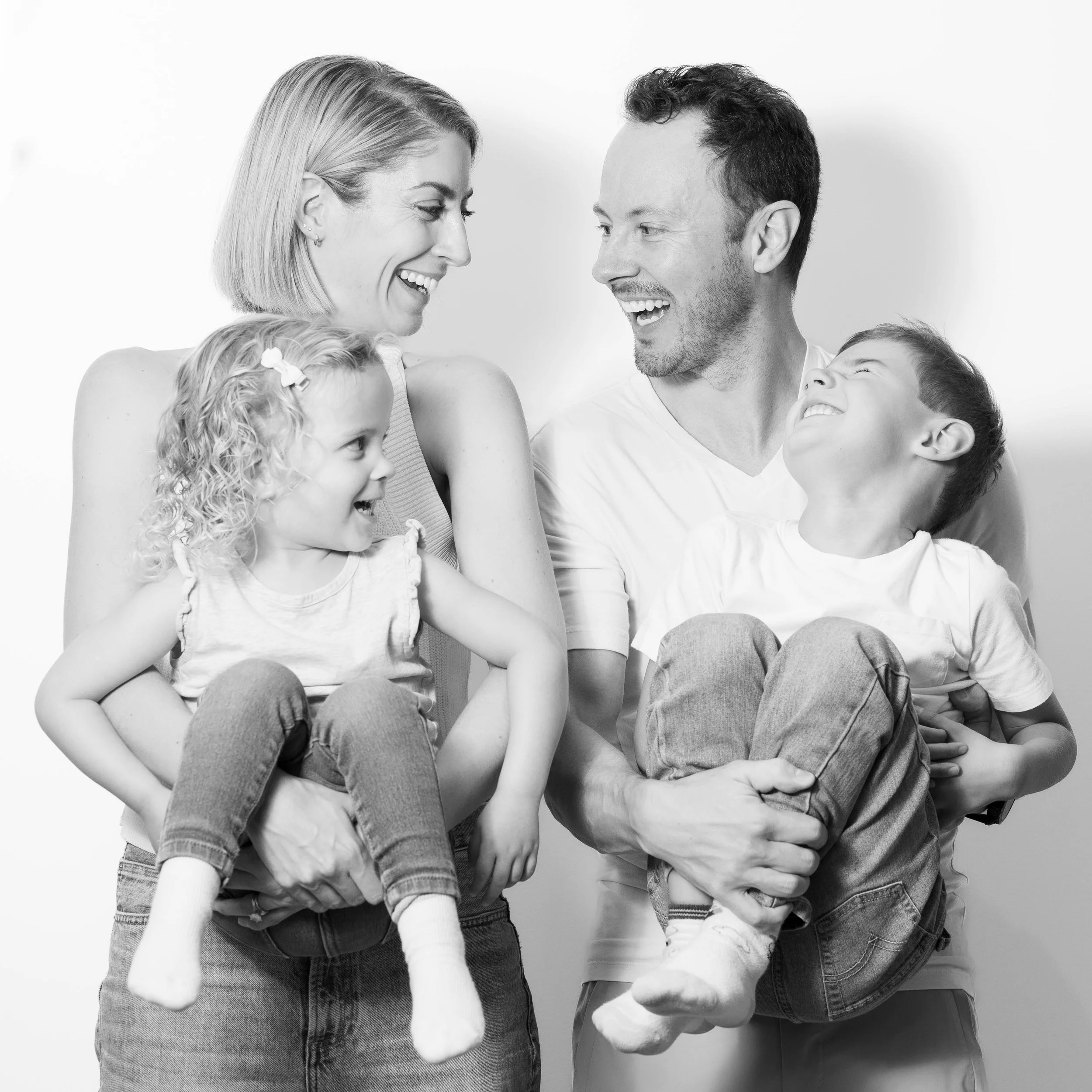 A happy family of four with two children, a mother, father, a young girl, and a young boy, all smiling and looking at each other in a playful pose, black and white photo.
