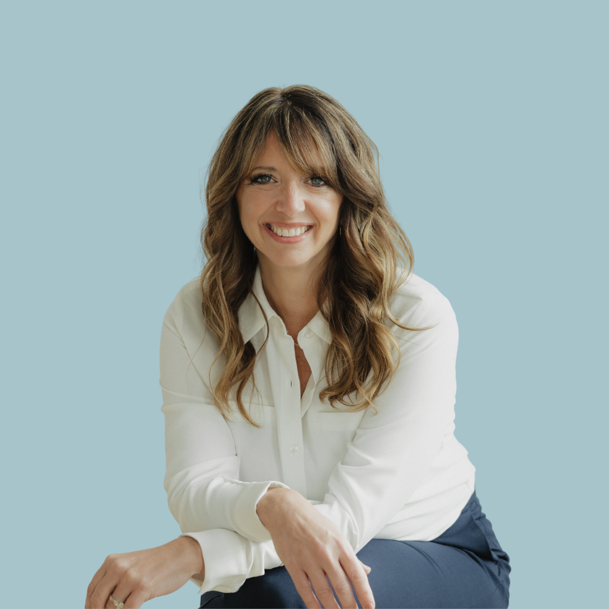 A woman with long wavy brown hair and a white shirt, smiling, sitting against a light blue background.