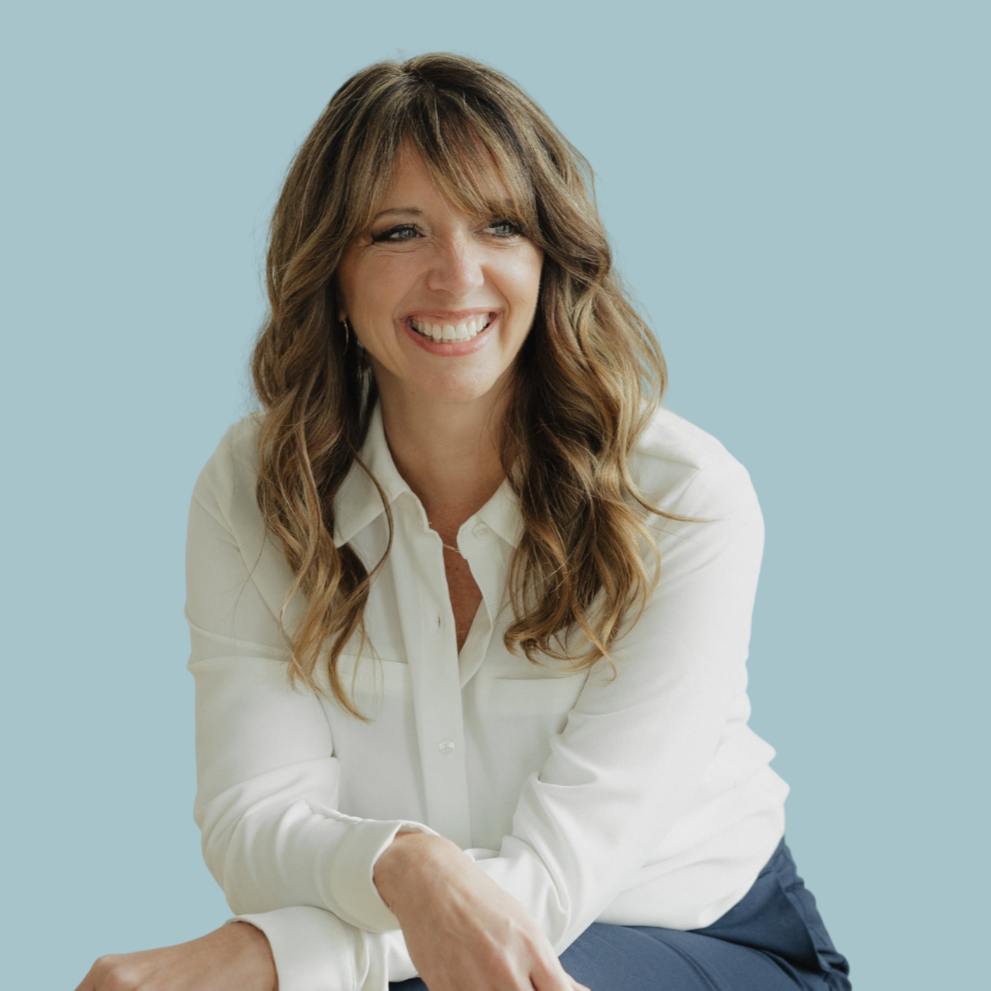 A woman with long wavy brown hair, smiling, wearing a white shirt and dark pants, sitting with one arm resting on her knee, against a light blue background.