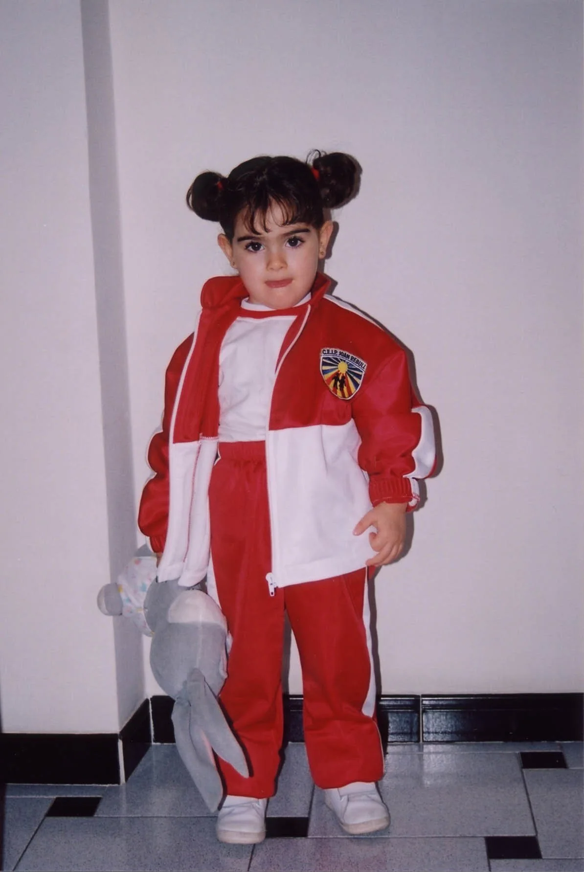 Niña vestida con uniforme de fútbol en blanco y rojo, sosteniendo un peluche de delfín, de pie en el suelo de baldosas negras y blancas junto a una pared blanca.