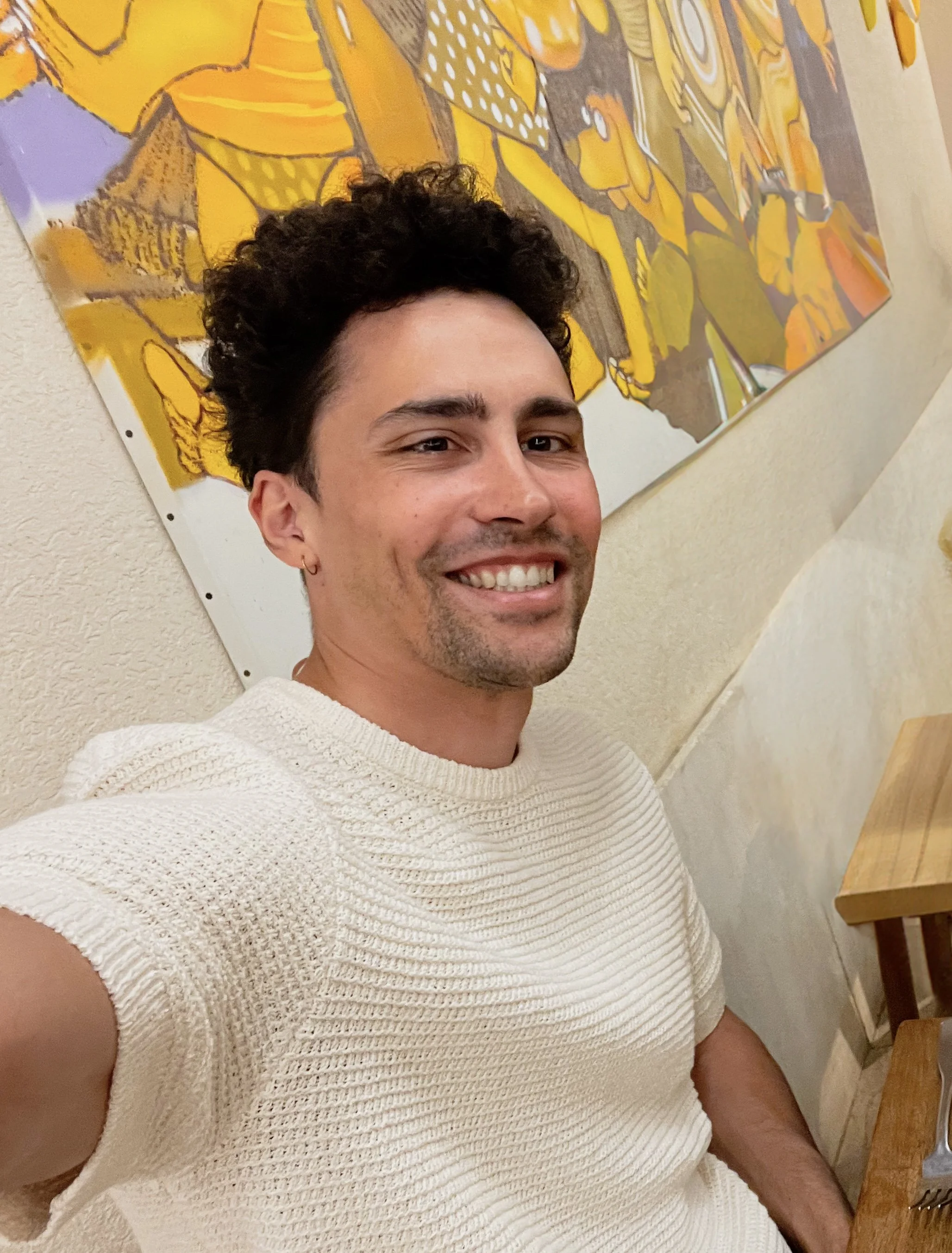 A man with dark curly hair and a small earring in his left ear, smiling and taking a selfie. He is wearing a cream-colored, textured short-sleeve sweater. Behind him, there is a colorful yellow, orange, and purple abstract art on a wall.