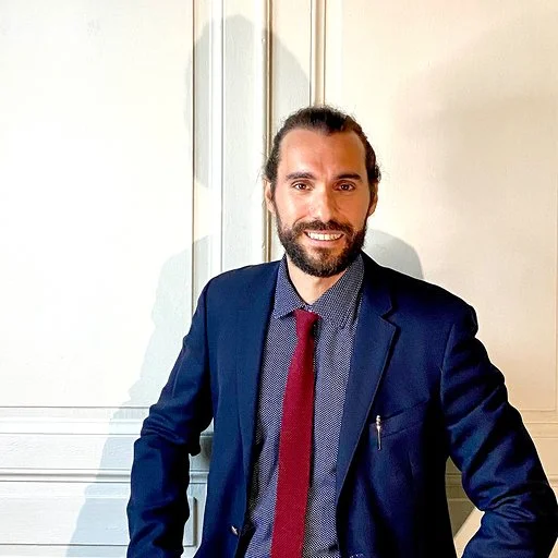 A man with a beard and long hair tied back, wearing a navy blue blazer, a blue dress shirt, and a red tie, standing and smiling indoors in front of white-paneled walls.