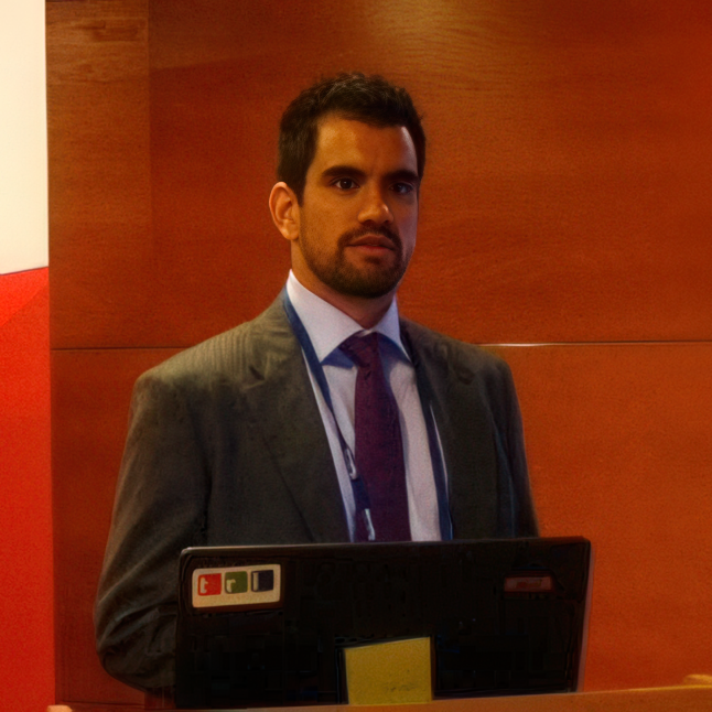 A man with dark hair, beard, dressed in a gray suit, white shirt, and purple tie standing in front of a wooden wall, with a laptop in front of him.
