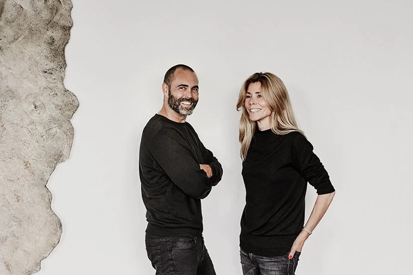 A man and a woman standing against a plain white wall with a textured section on the left side, both smiling and dressed in black tops.