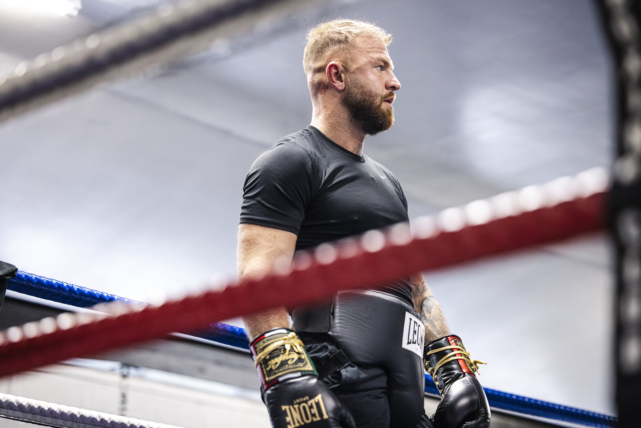 A male boxer with a beard and tattoos on his left arm stands inside a boxing ring, wearing black boxing gloves, black shorts, and a black t-shirt, appearing focused during a training session.