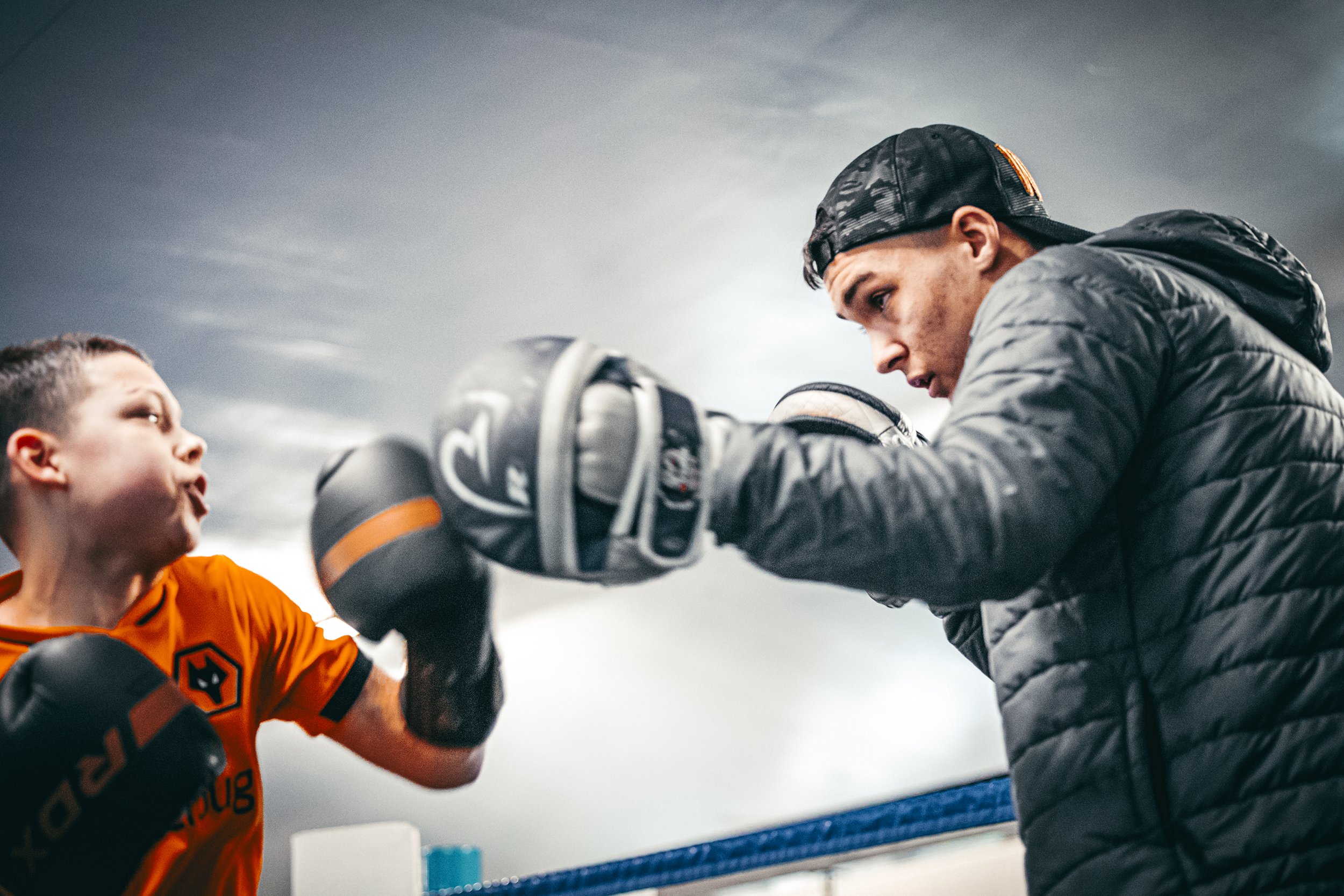A young boy training in boxing with a coach inside a boxing ring during daytime. The boy is wearing an orange boxing shirt and black boxing gloves, and the coach is wearing a black quilted jacket, black cap backward, and boxing gloves, focusing inten