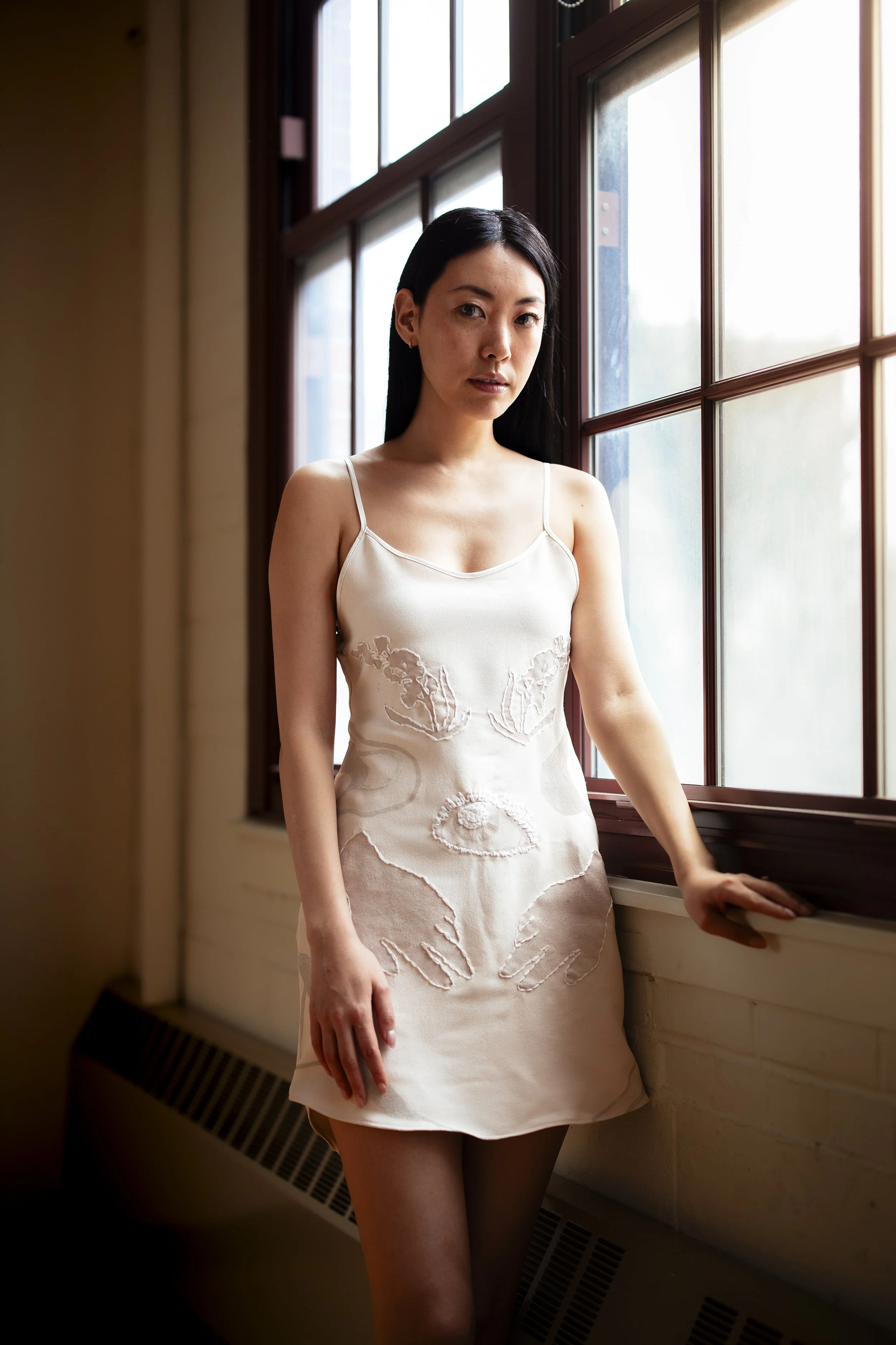 A woman with long black hair wearing a cream-colored spaghetti strap dress with floral embroidery, standing by a window with wooden frames, with her hand resting on the windowsill.