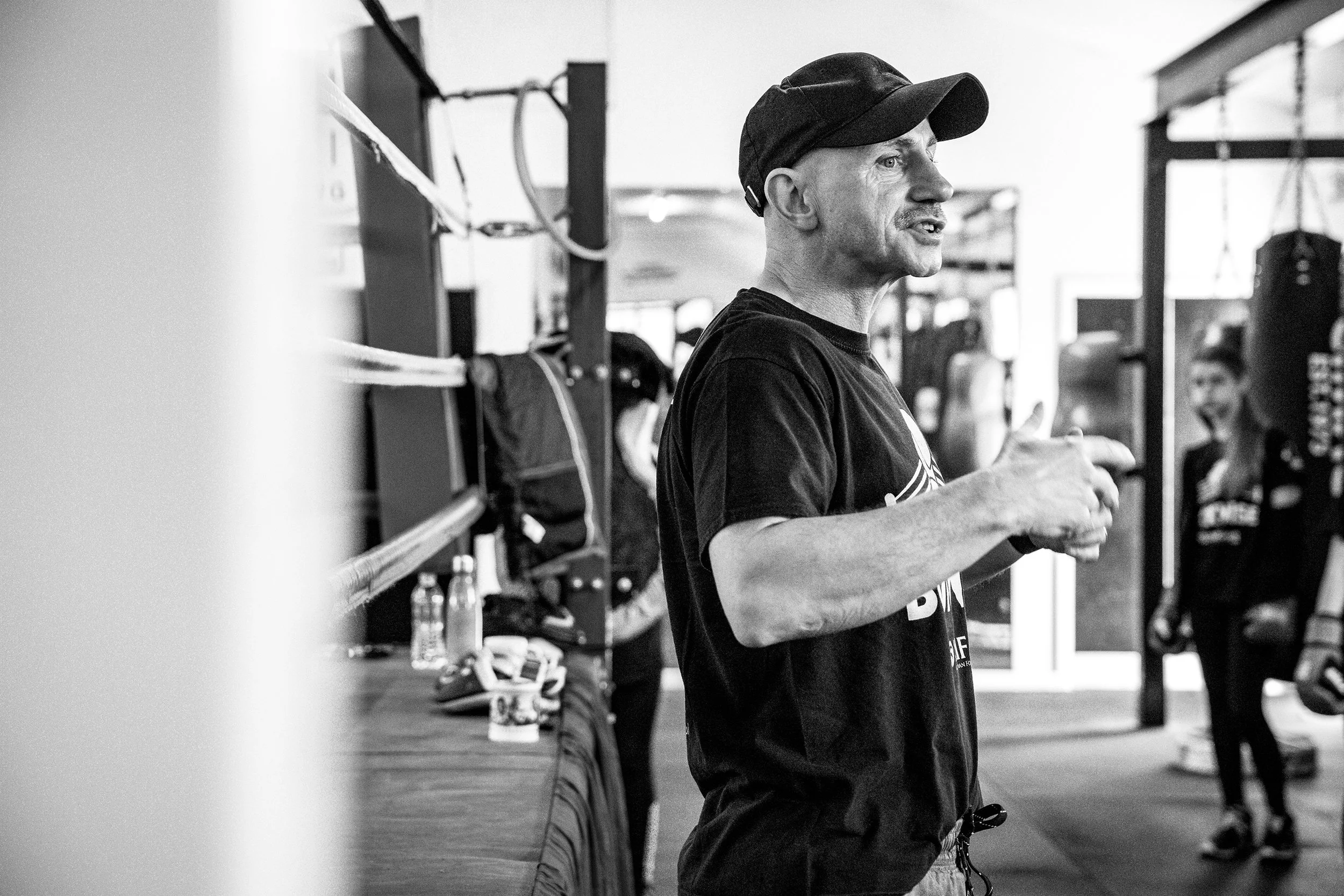 A man in a black cap and black t-shirt talking in a gym, with other people in the background participating in fitness activities.