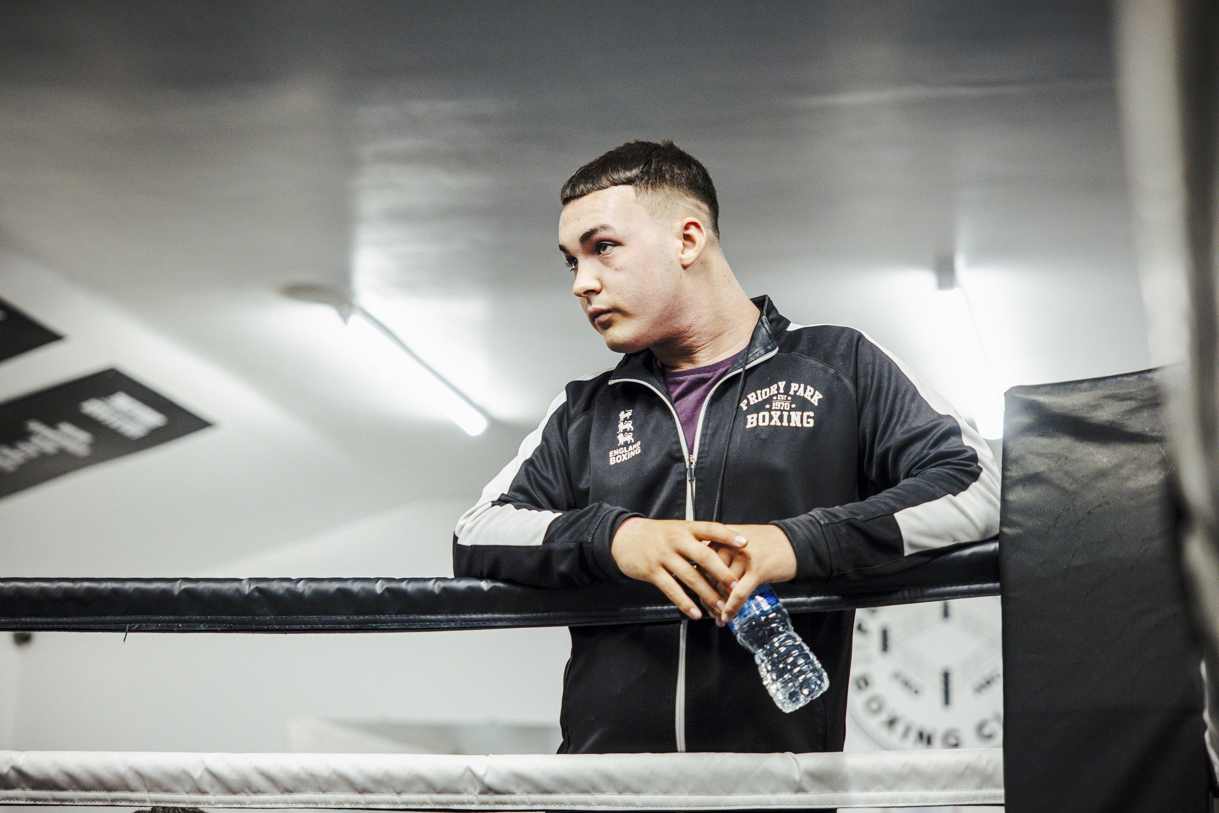 A young man leaning on a boxing ring corner, holding a water bottle, wearing a jacket with 'Friday Park Boxing' text, inside a gym with white walls and fluorescent lighting.
