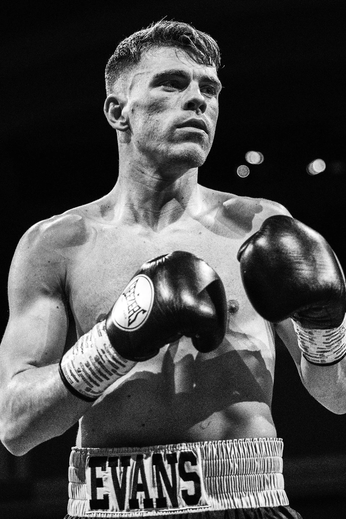 Black and white photo of a male boxer with gloves, shorts, and a focused expression, standing in a boxing ring.