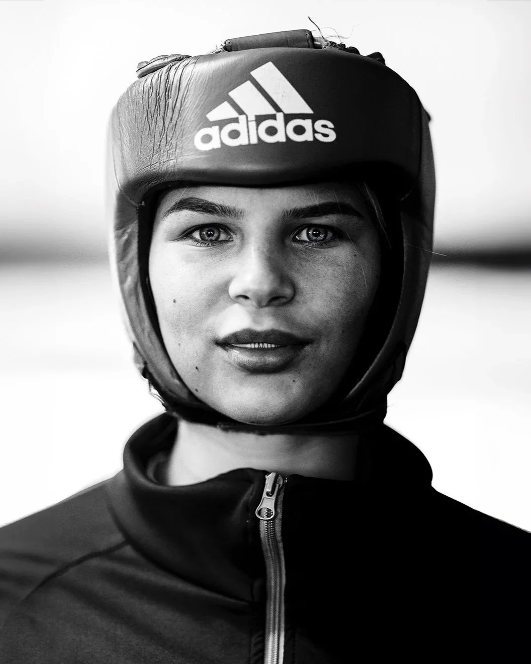 A young woman with light eyes and freckles wearing a black Adidas helmet and black jacket, looking at the camera in black and white.