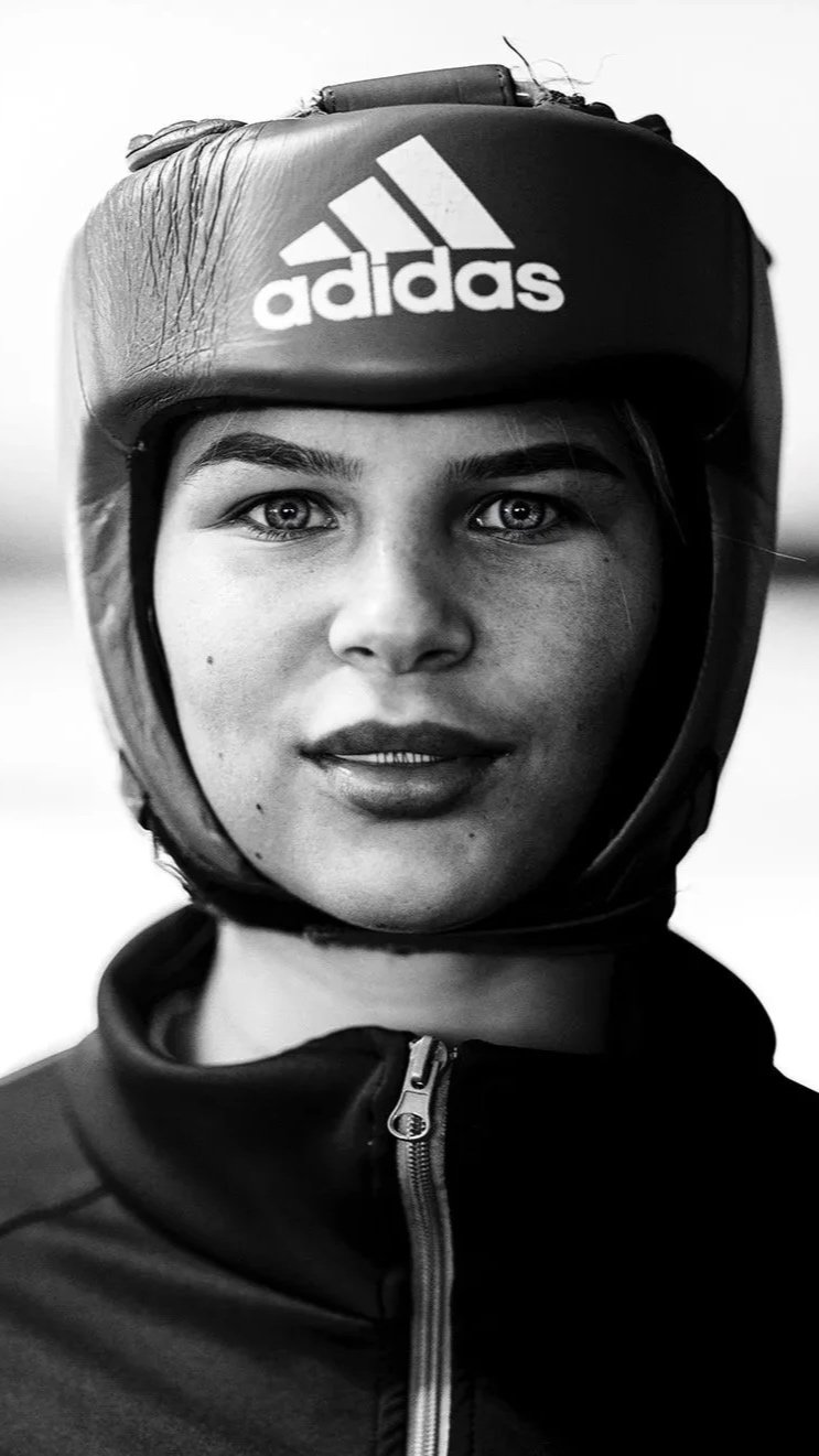 A woman with light-colored eyes wearing a black Adidas helmet with a zipper jacket.