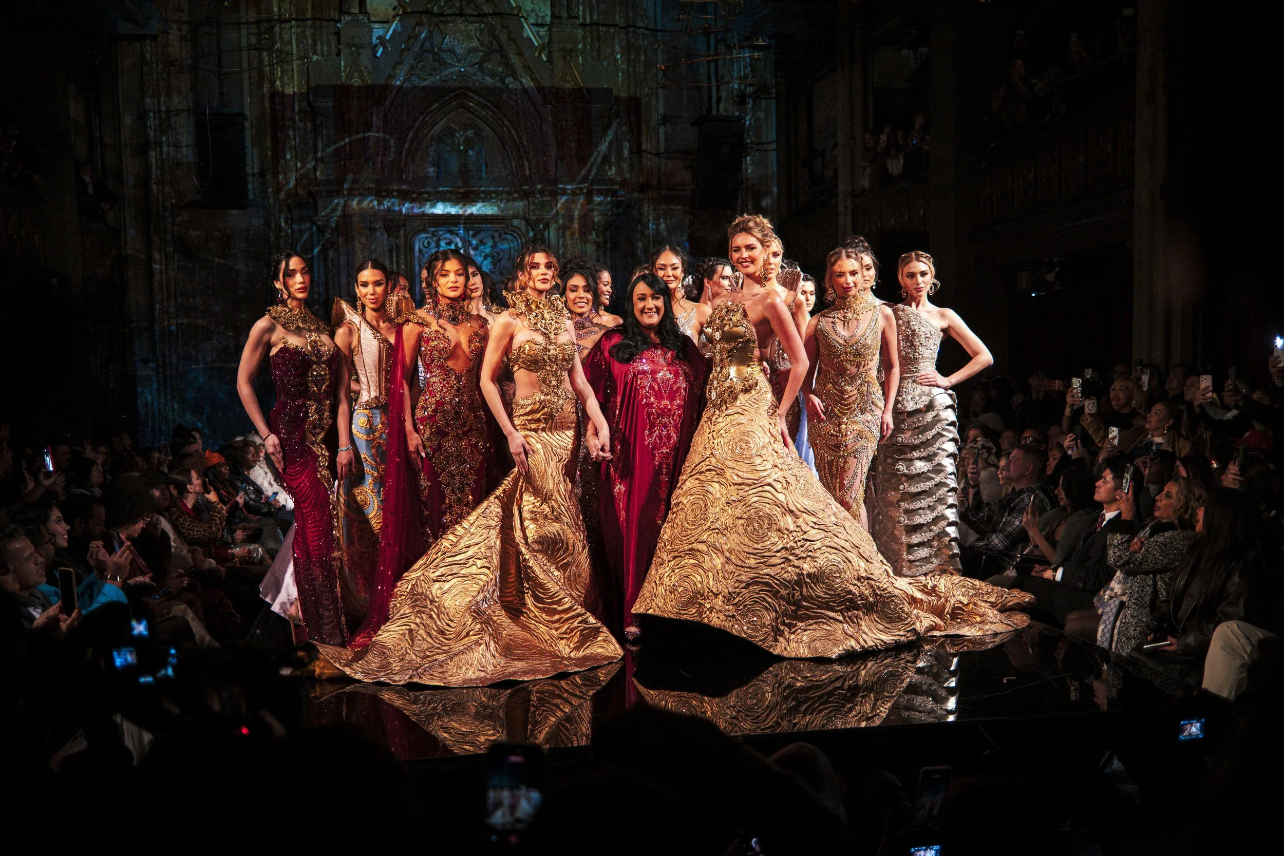 Group of women in elaborate, colorful gowns walking on a runway during a fashion show with an audience taking photos.