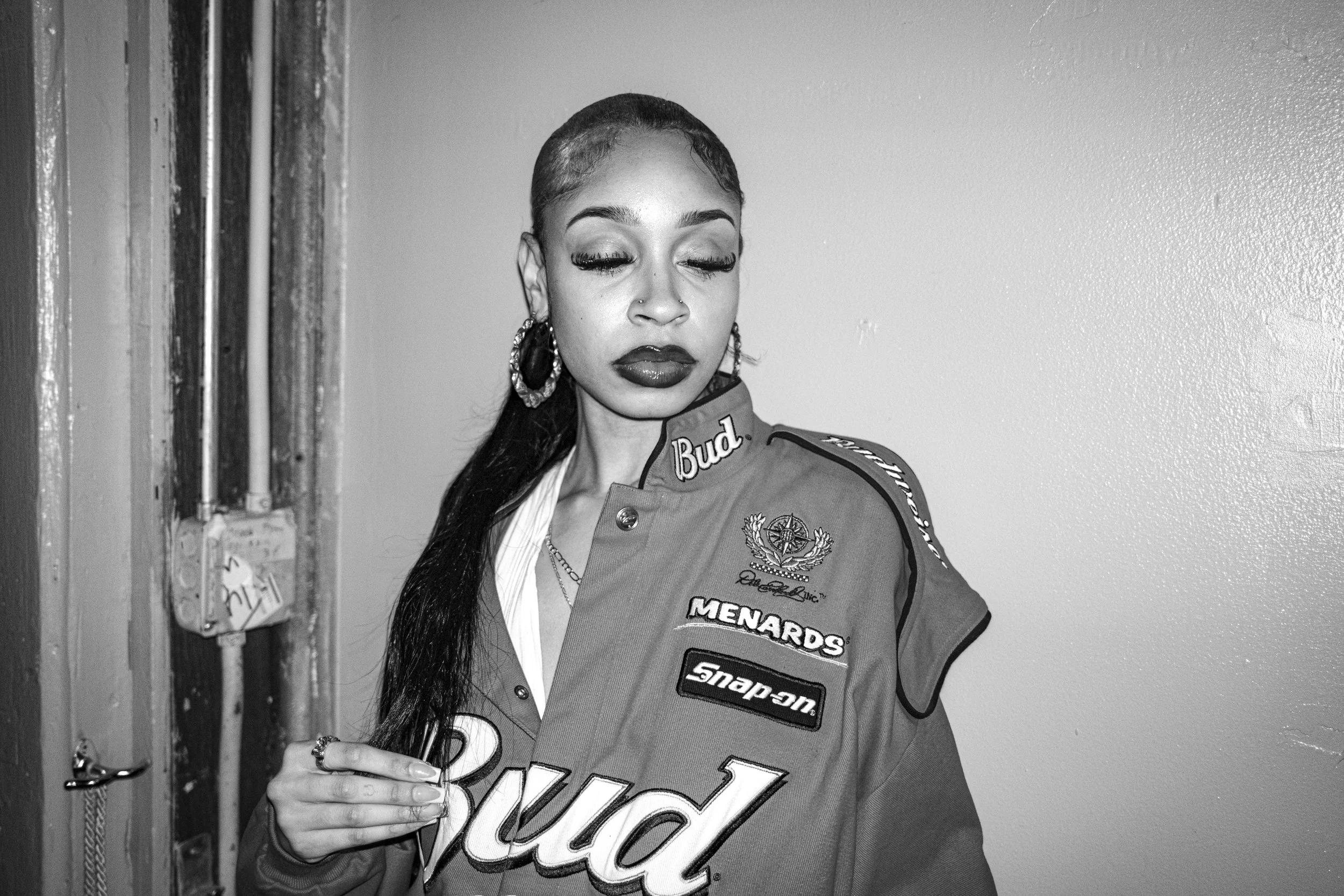 Black and white photo of a woman with long hair styled close to her scalp, wearing large hoop earrings, and a racing jacket with sponsor patches. She has her eyes closed and is holding a phone in her right hand.
