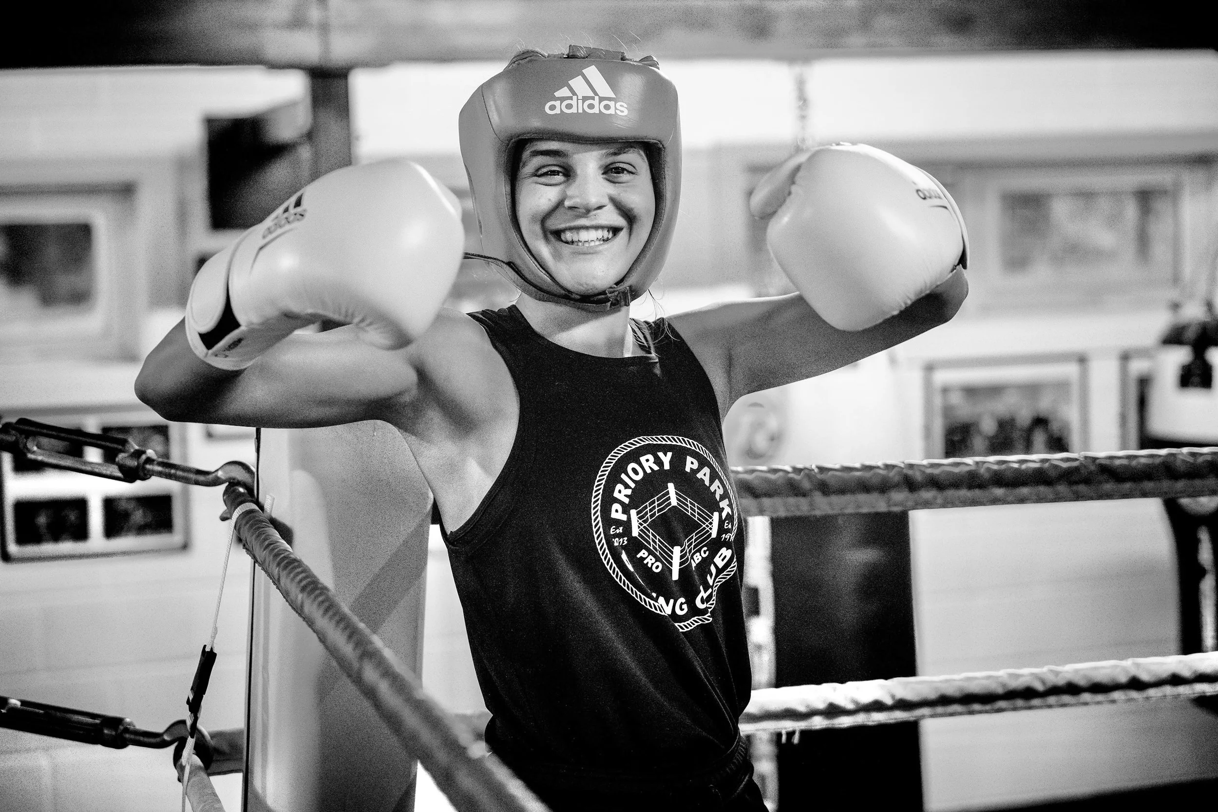 A young woman boxer smiling, wearing a padded helmet and boxing gloves, standing in a boxing ring with her arms raised in victory.