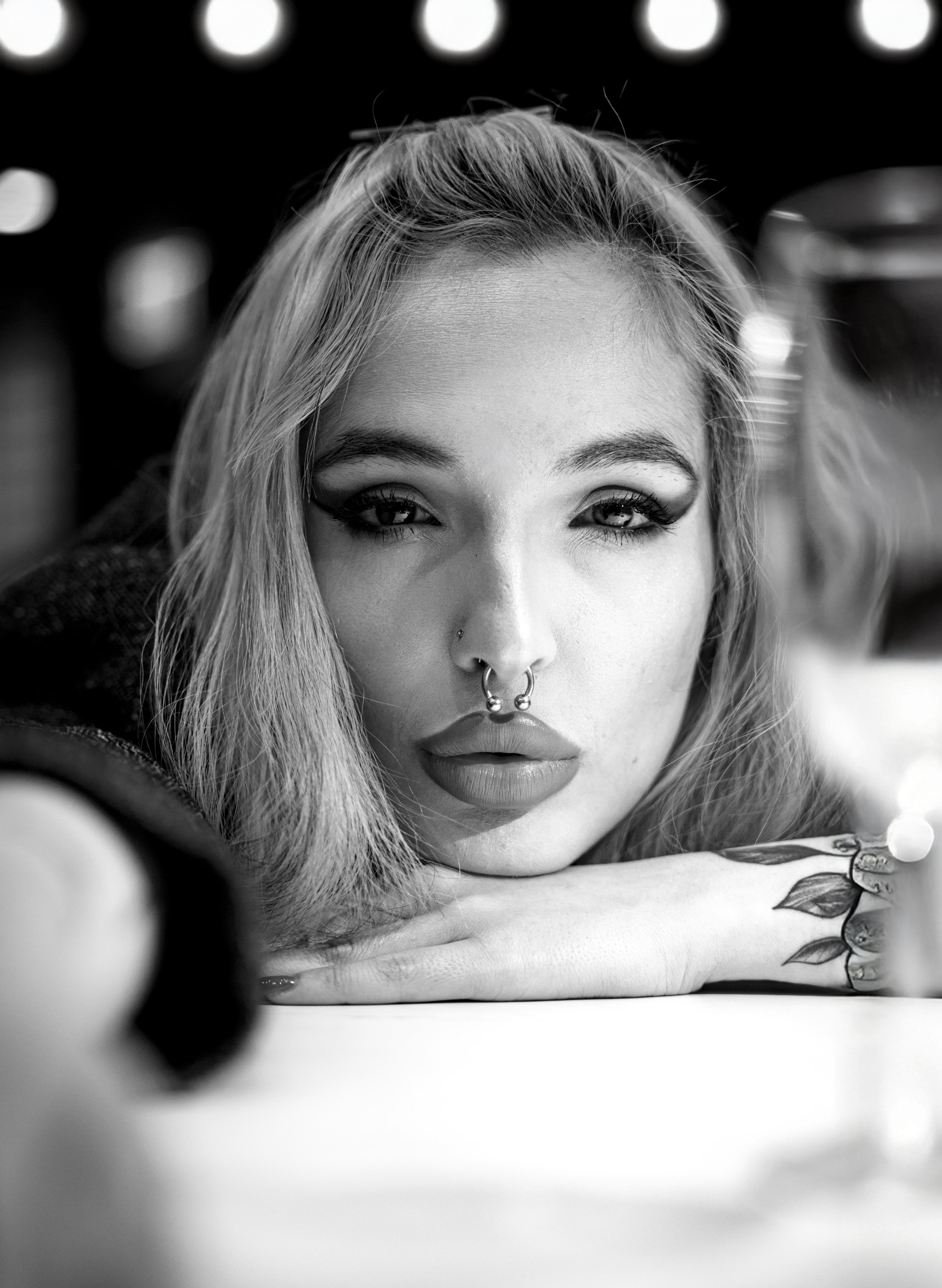 Black and white close-up of a woman with light hair, piercings, makeup, and tattoos resting her chin on her crossed arms, with a blurred background.