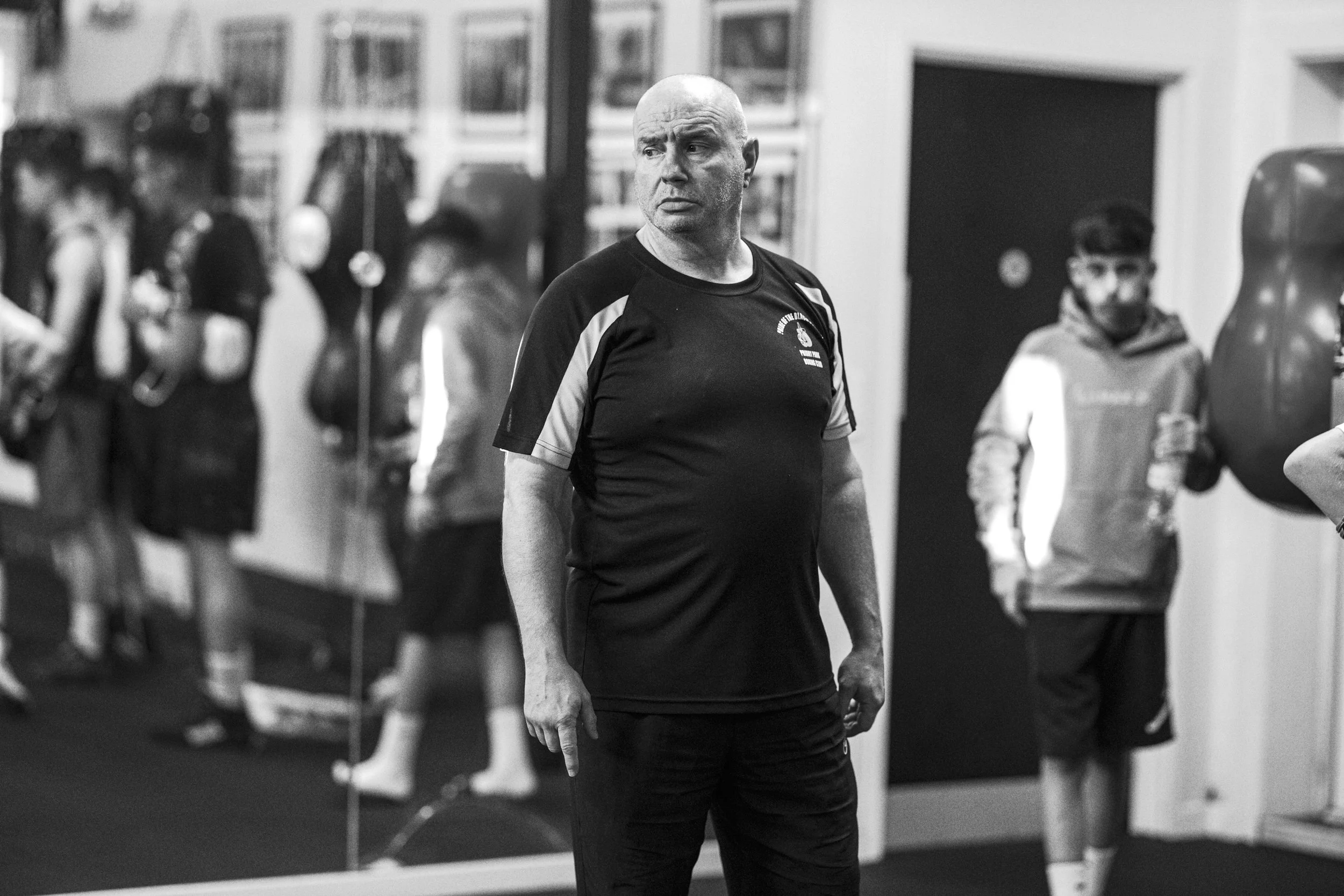 A man standing in a gym with other people in the background. The man appears to be a coach or trainer, wearing sportswear.