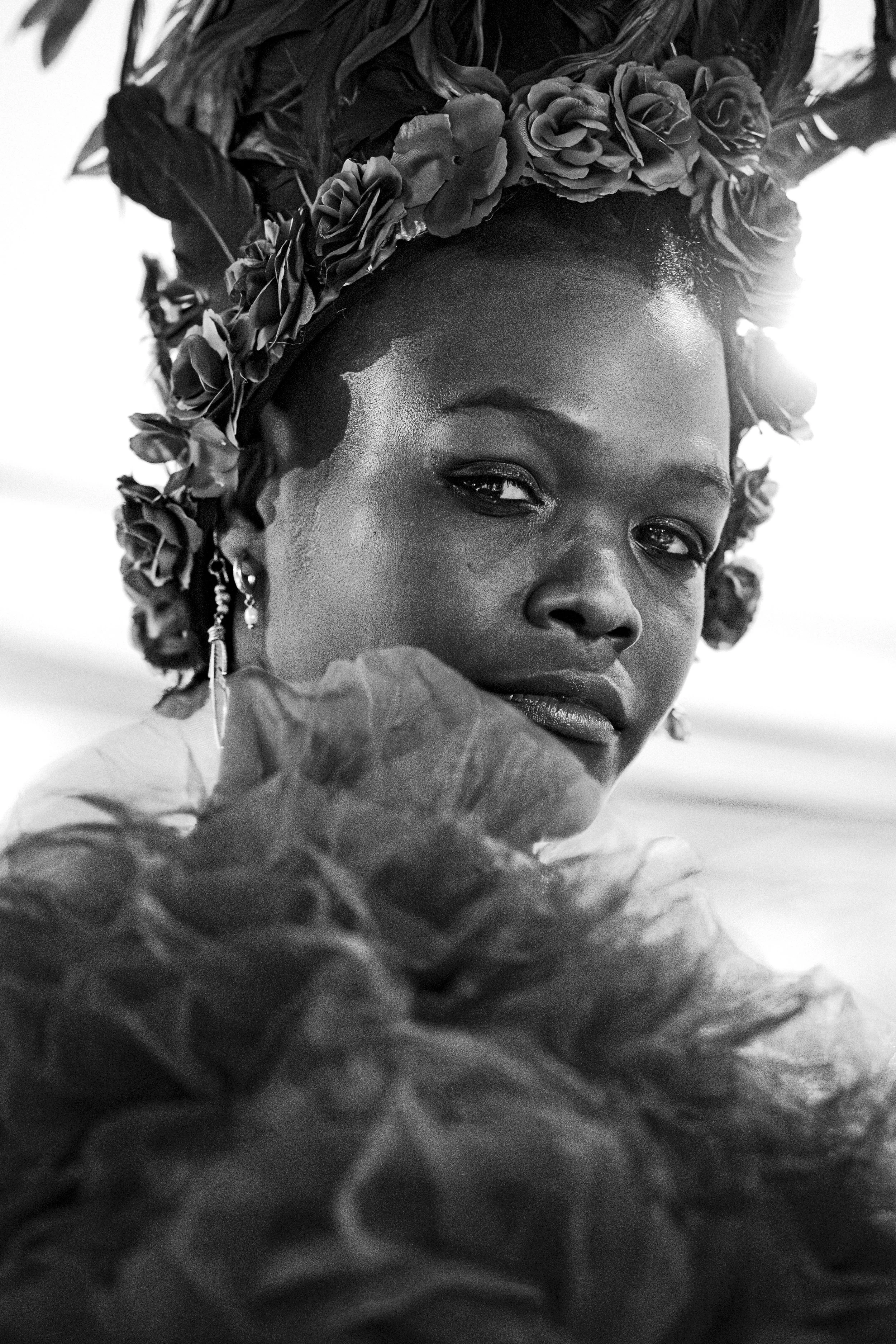A black and white portrait of a woman wearing a floral headpiece and earrings, with a serious expression and textured clothing.