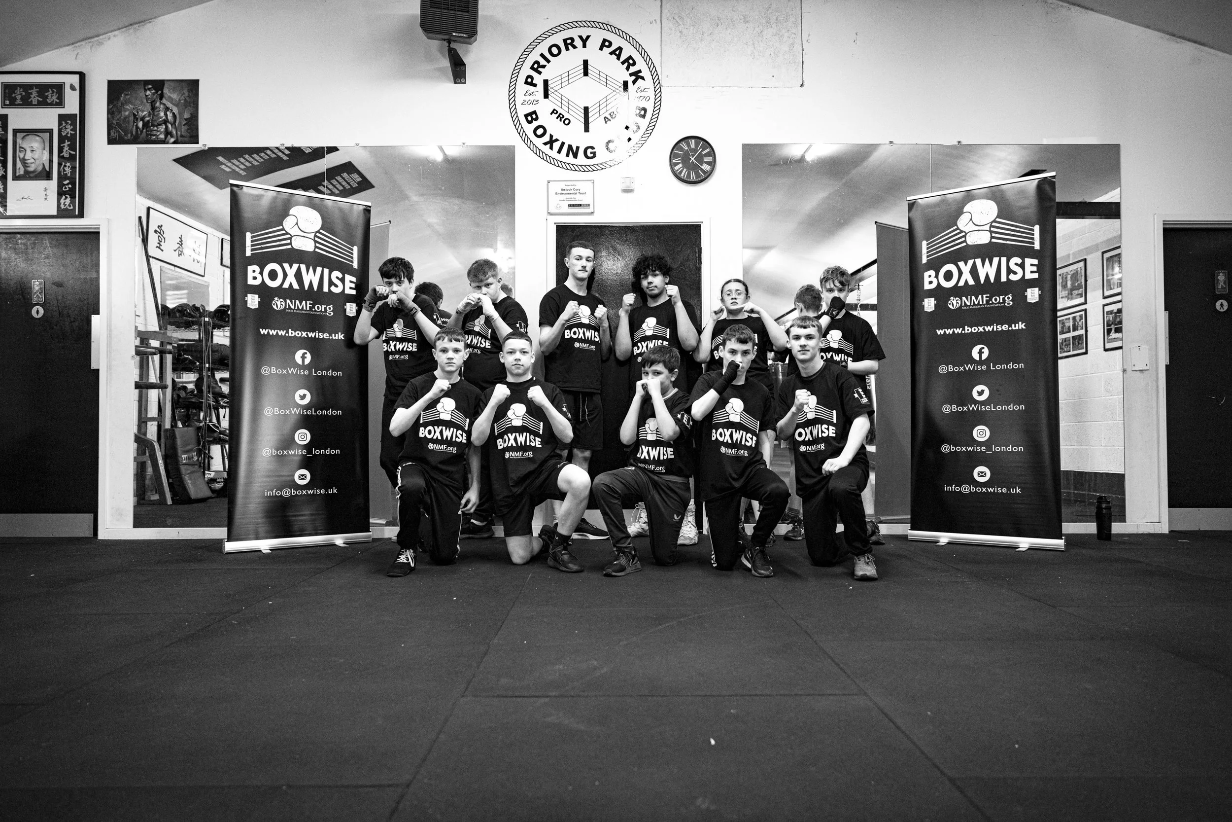 A group of young boxers in T-shirts with 'BoxWise' logo posing in a boxing gym, standing and kneeling with fists raised. They are in front of promotional banners for BoxWise, with a boxing ring logo and social media handles.