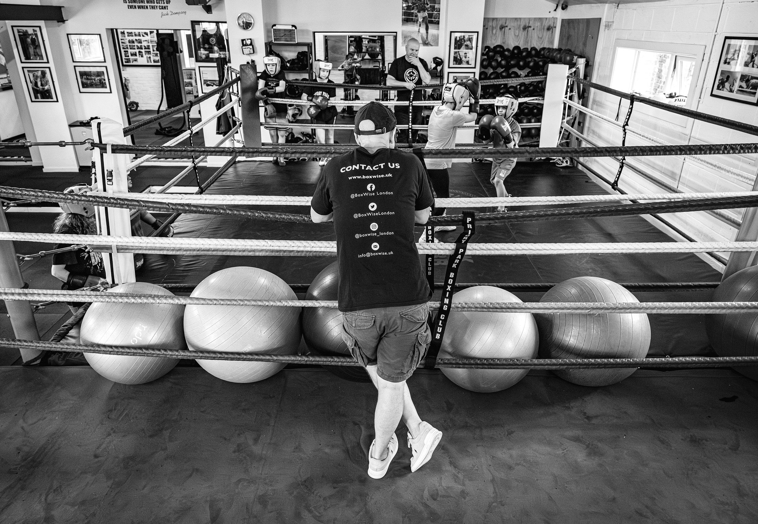 A boxing gym with a boxing ring where young children are practicing boxing under supervision. A man, possibly a coach, stands with his back to the camera observing the gym. The gym has large exercise balls at the front, and walls decorated with frame
