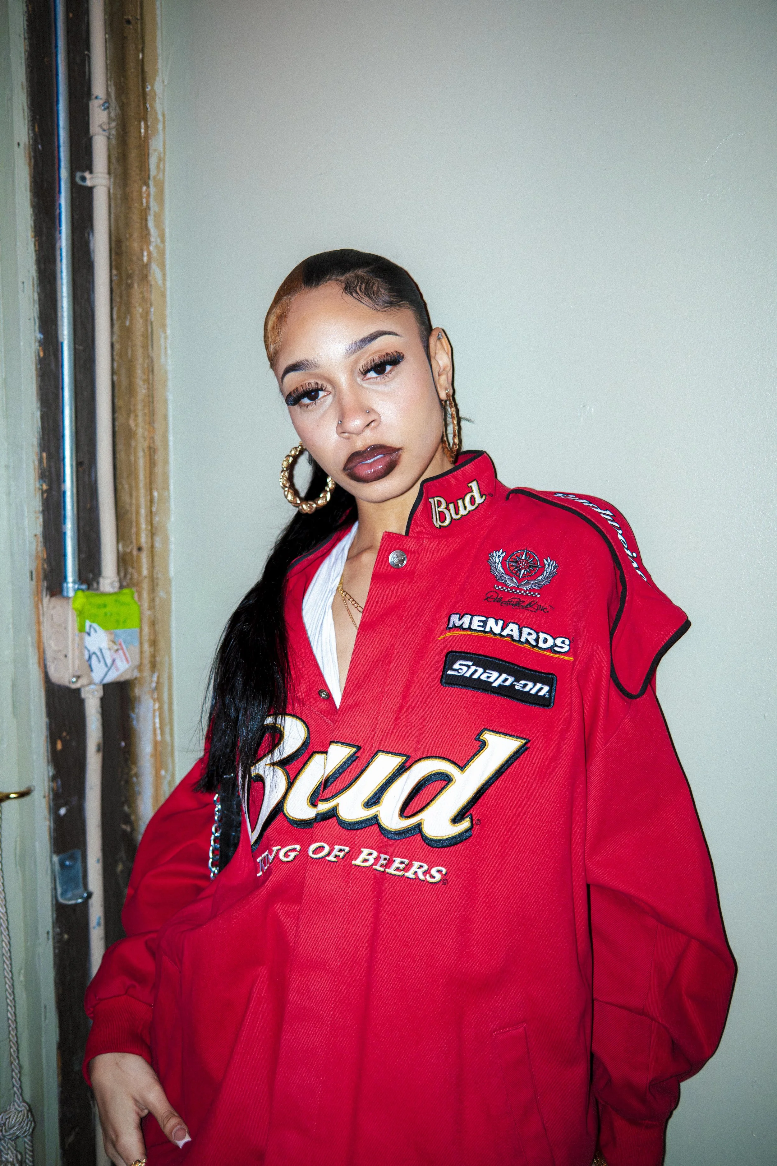 A woman with long black hair, hoop earrings, and dark makeup wearing a red Budweiser jacket with patches and logos, standing indoors near a partially exposed wall and electrical wiring.
