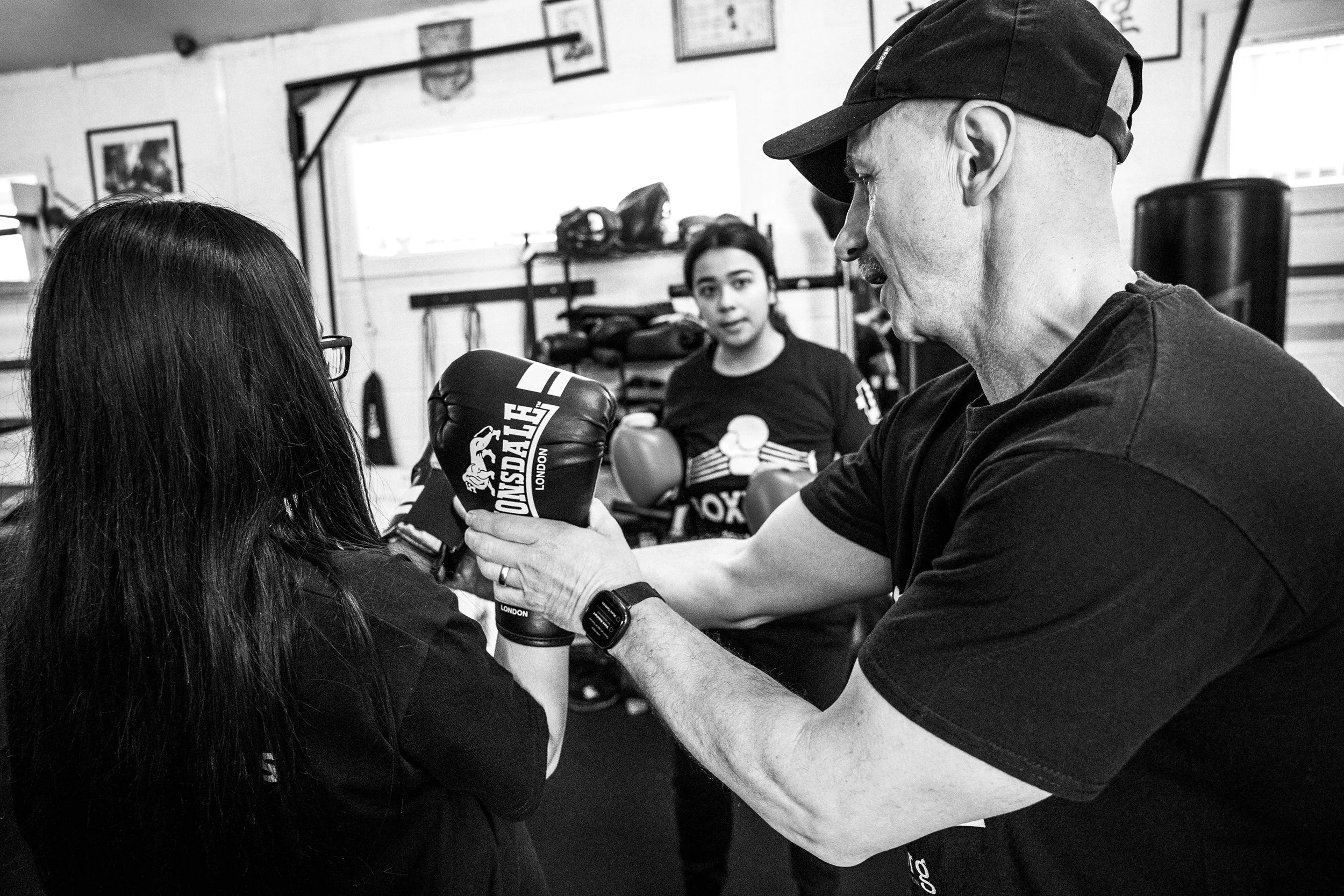A boxing instructor demonstrates a technique to a female student during a training session in a gym. Another woman watches in the background.