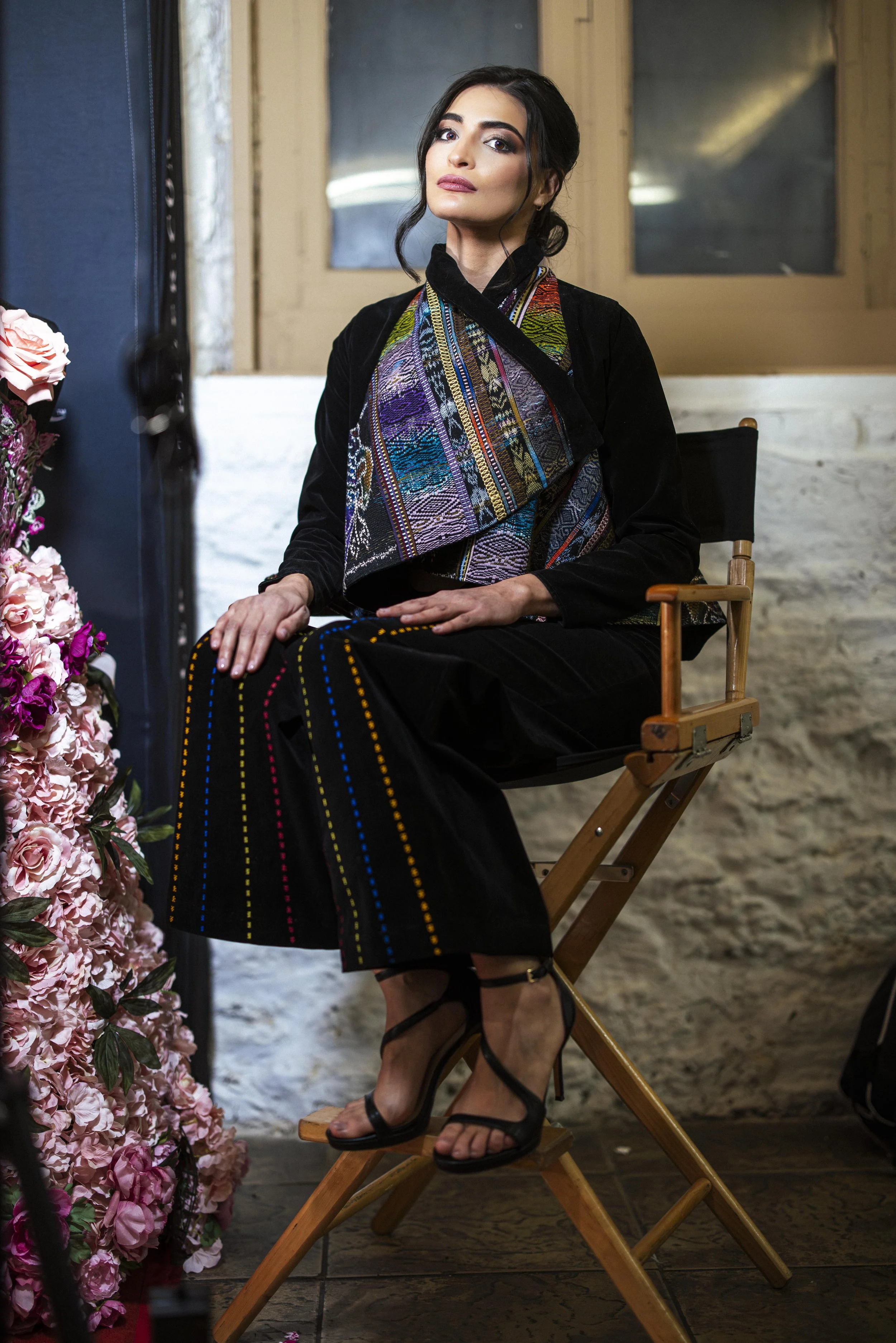 A woman with dark hair in an updo sitting on a wooden chair, wearing a colorful embroidered shawl over black clothing, black high-heeled sandals, in front of a window with a floral arrangement to her left.