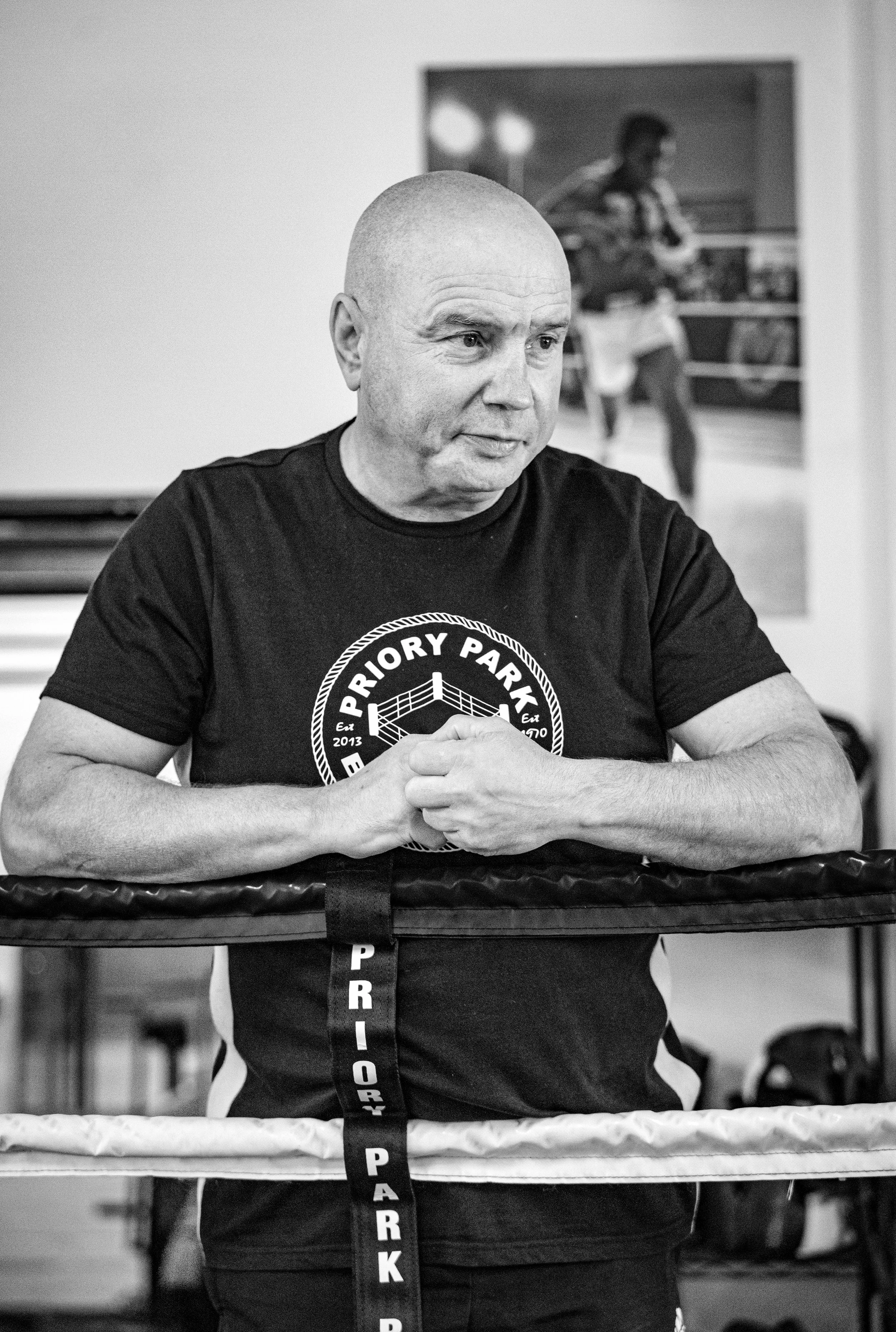 A bald man wearing a Priory Park t-shirt standing inside a boxing ring, with boxing ropes in front of him and a poster of a person playing guitar on the wall behind him.