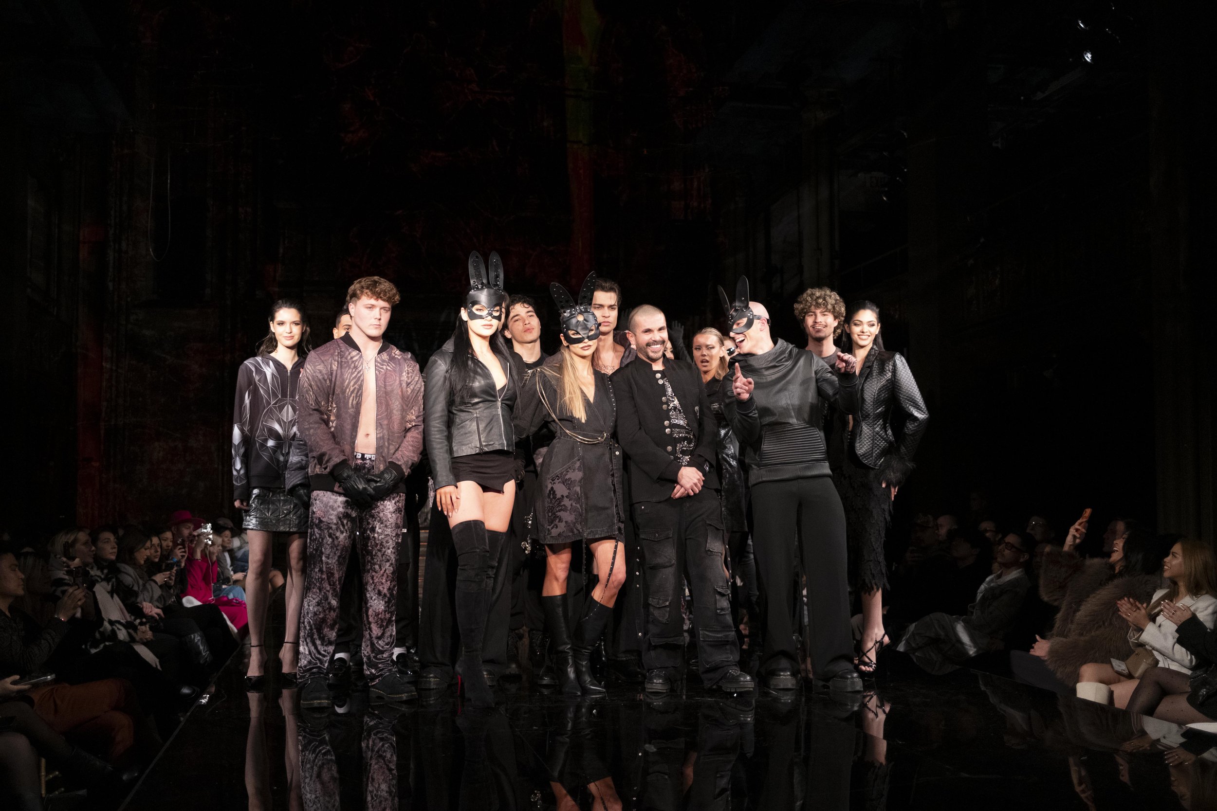 Models walking on a runway at a fashion show, dressed in dark, edgy clothing with some wearing black masks with bunny ears, while an audience watches.