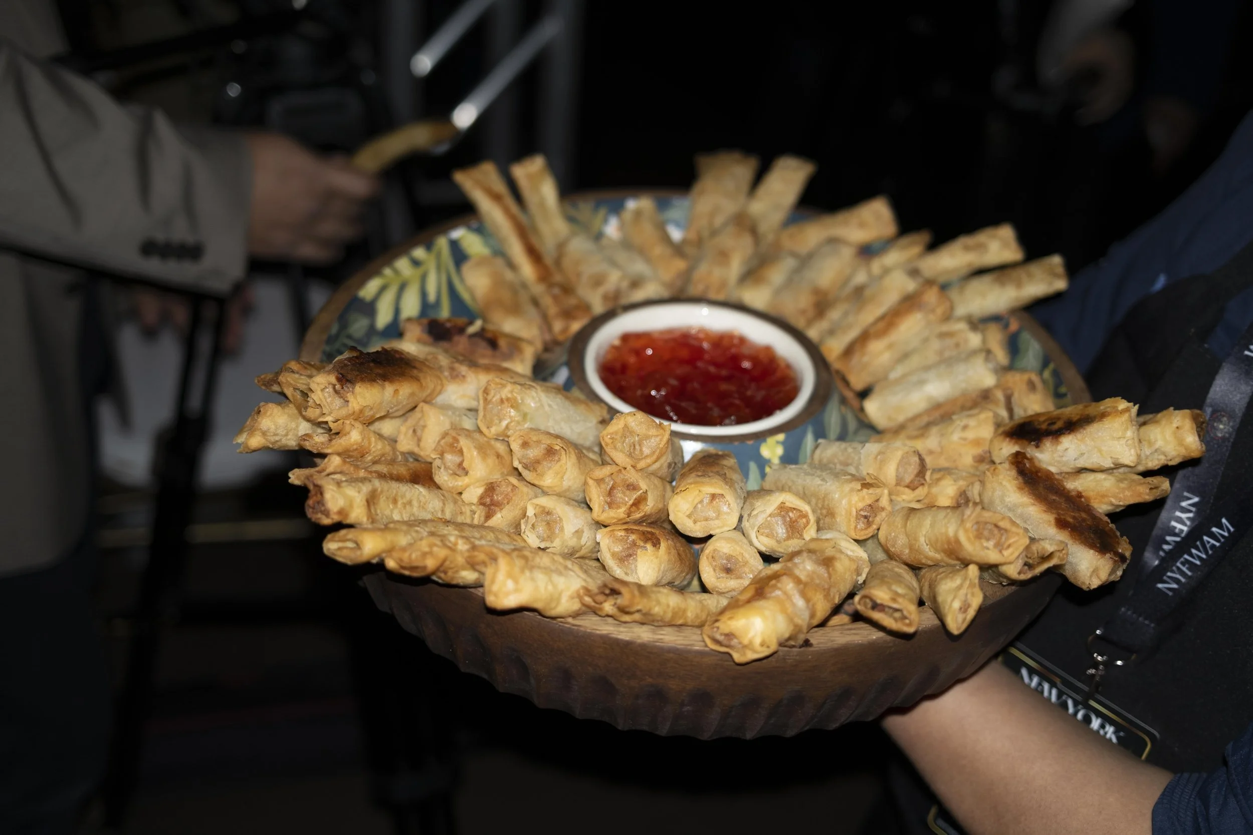 A person holds a wooden platter with fried spring rolls and a bowl of sweet chili sauce in the center.