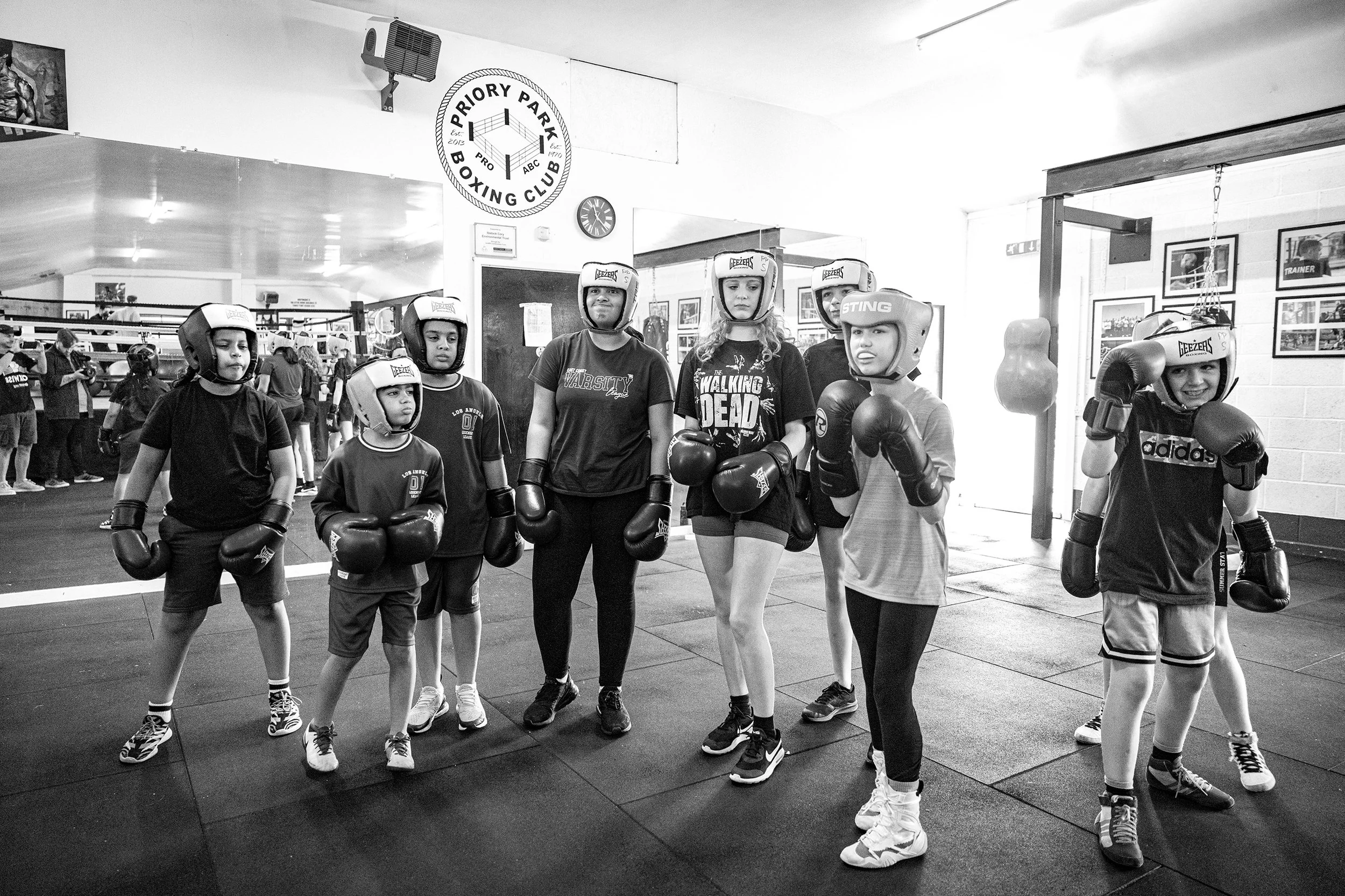 A group of children and a woman pose in a boxing gym, all wearing boxing helmets and gloves, in front of a boxing ring.