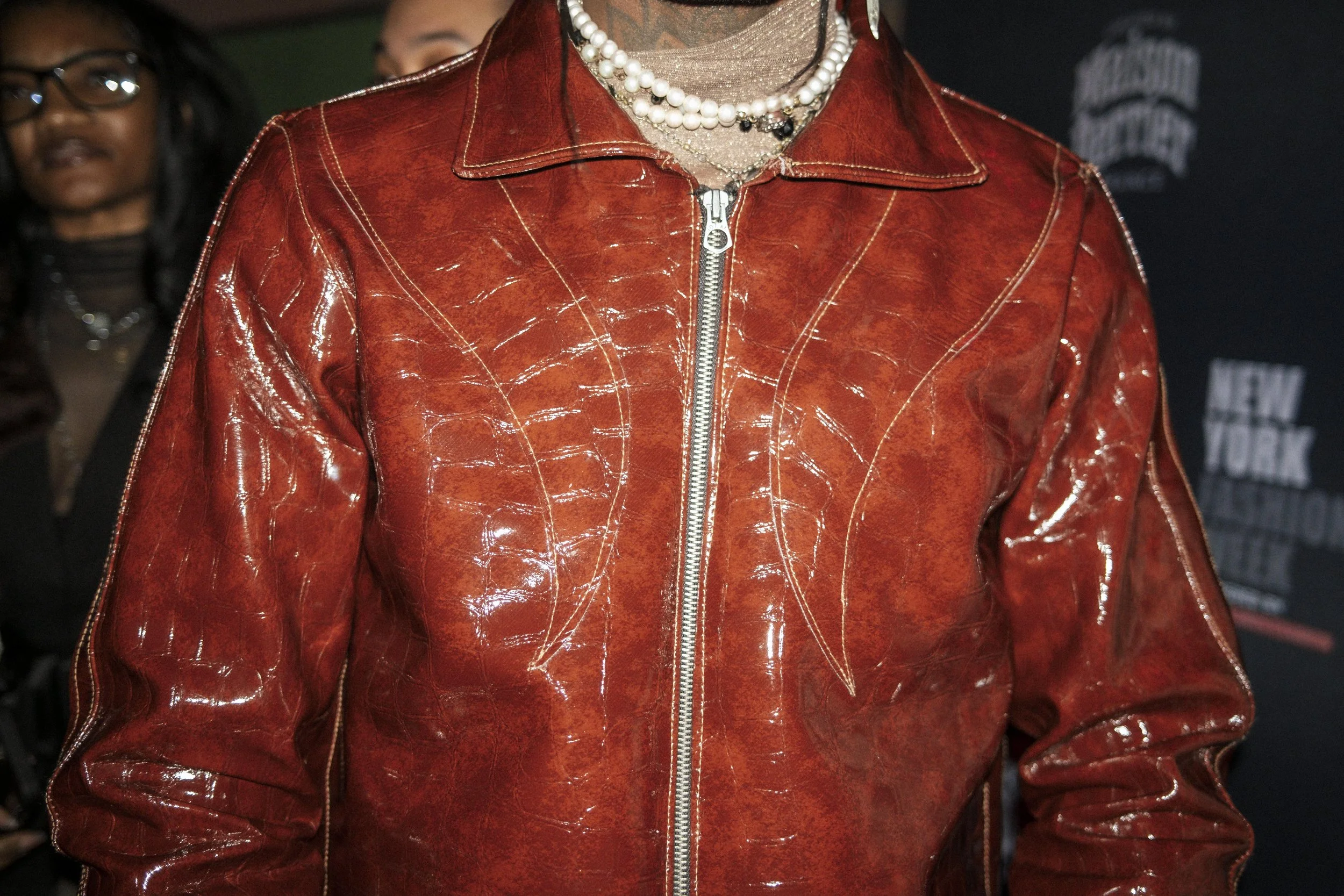 Close-up of person wearing a shiny red leather jacket with a silver zipper, layered pearl necklaces, and a top with a high neck in the background.
