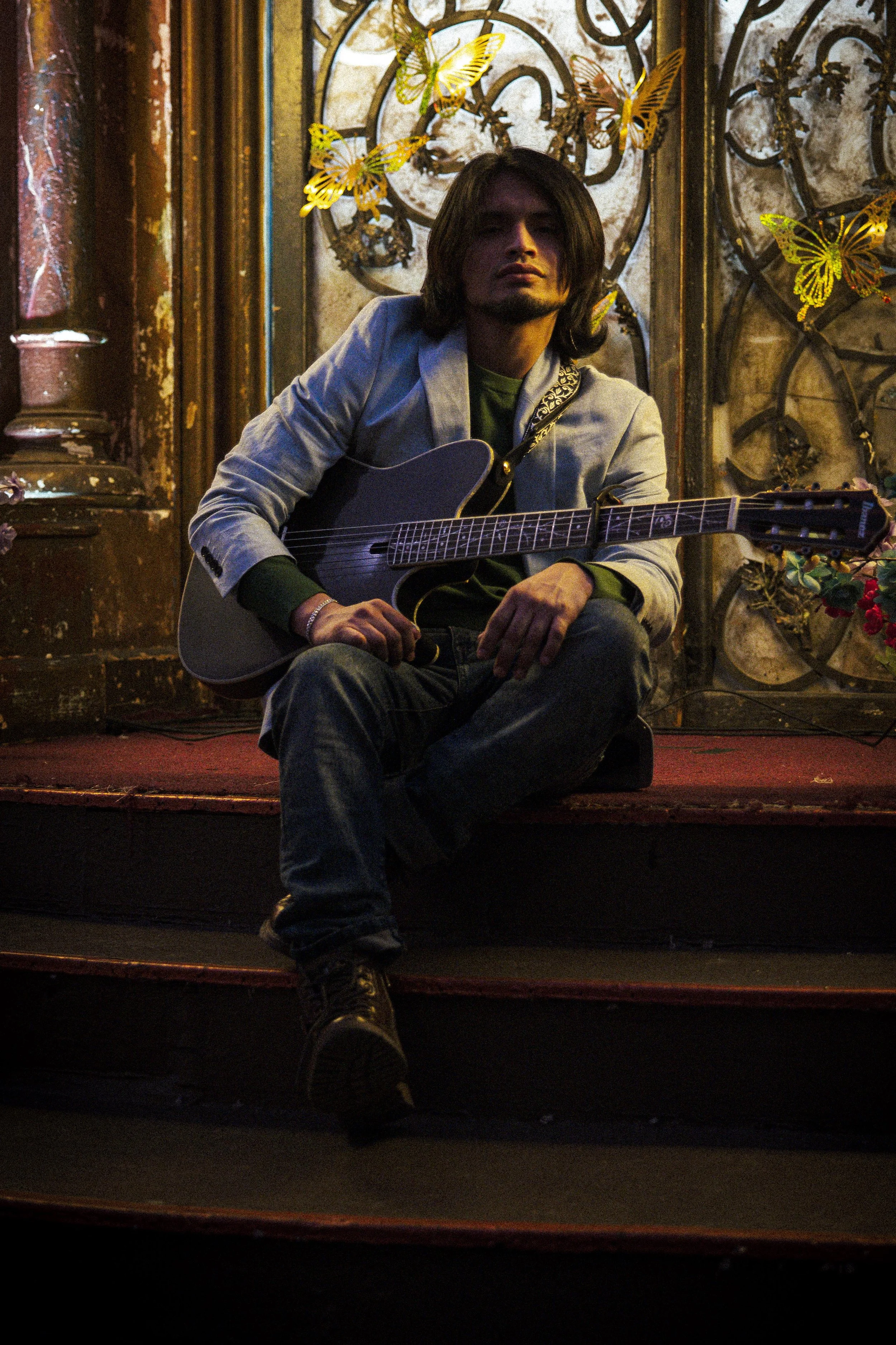 A young man with long hair and a light-colored blazer is sitting on stairs, holding an acoustic guitar. The background features a decorative wall with butterflies and floral designs.