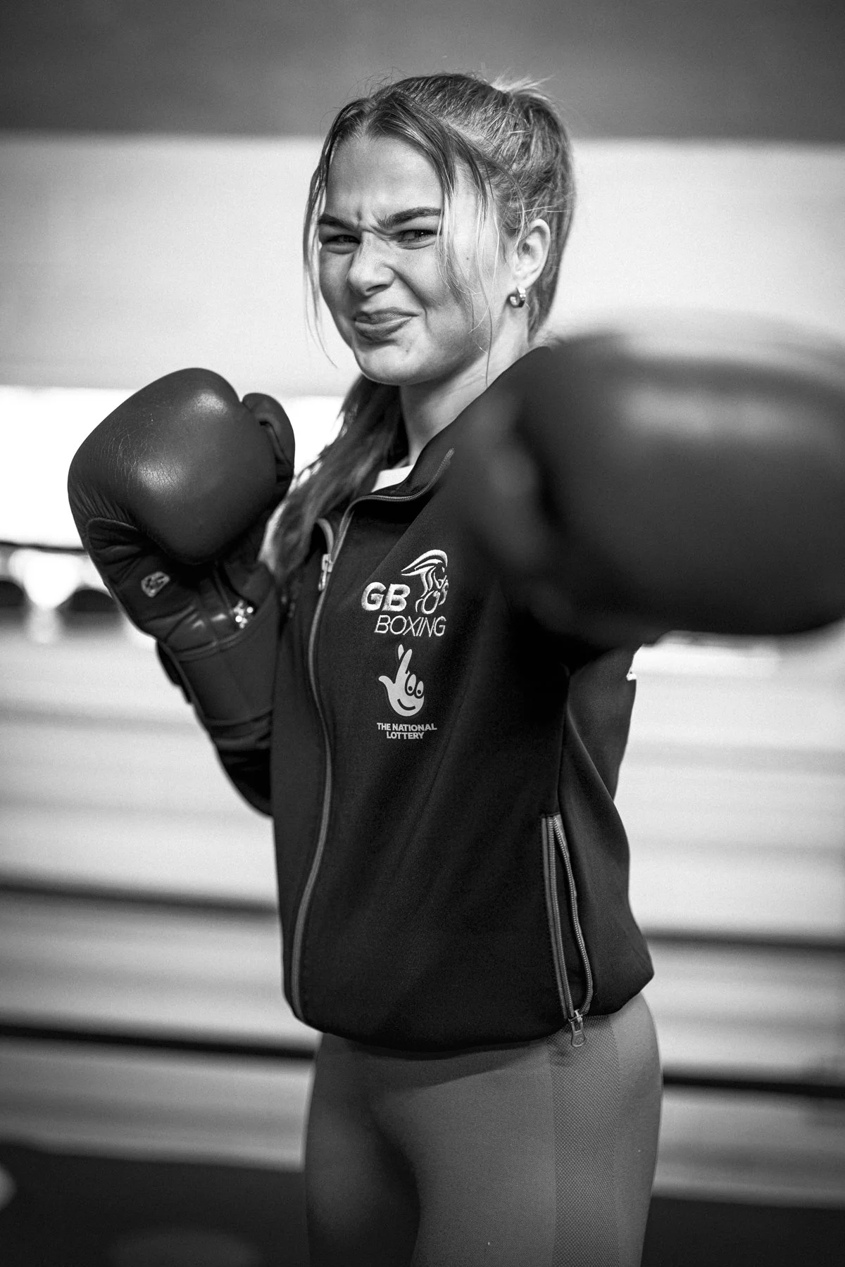 A woman wearing boxing gloves and a jacket with England boxing logo, making a playful face and pointing at the camera in a boxing gym.