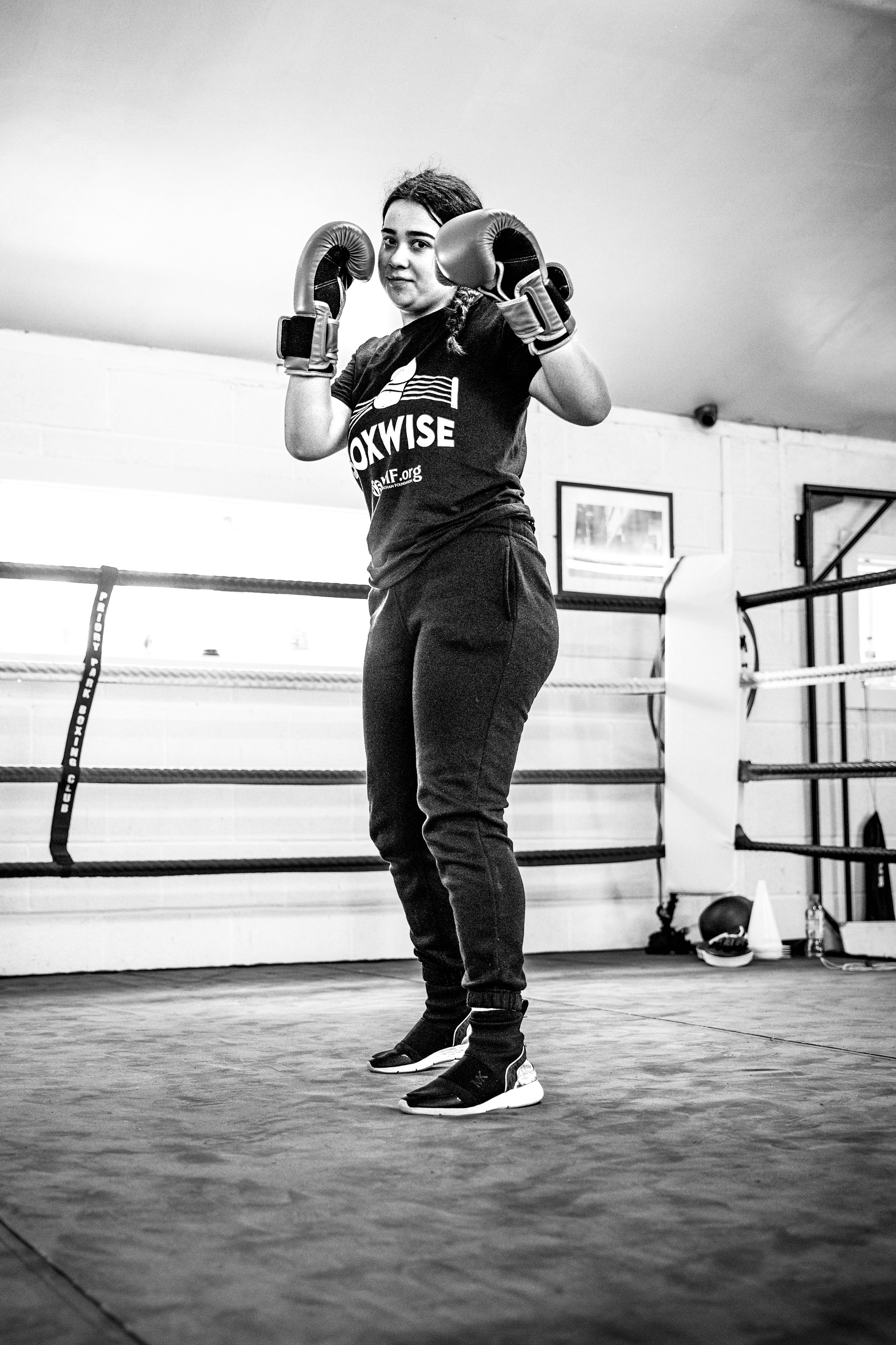 A woman standing in a boxing ring, wearing boxing gloves, a T-shirt, and athletic pants, posing confidently.