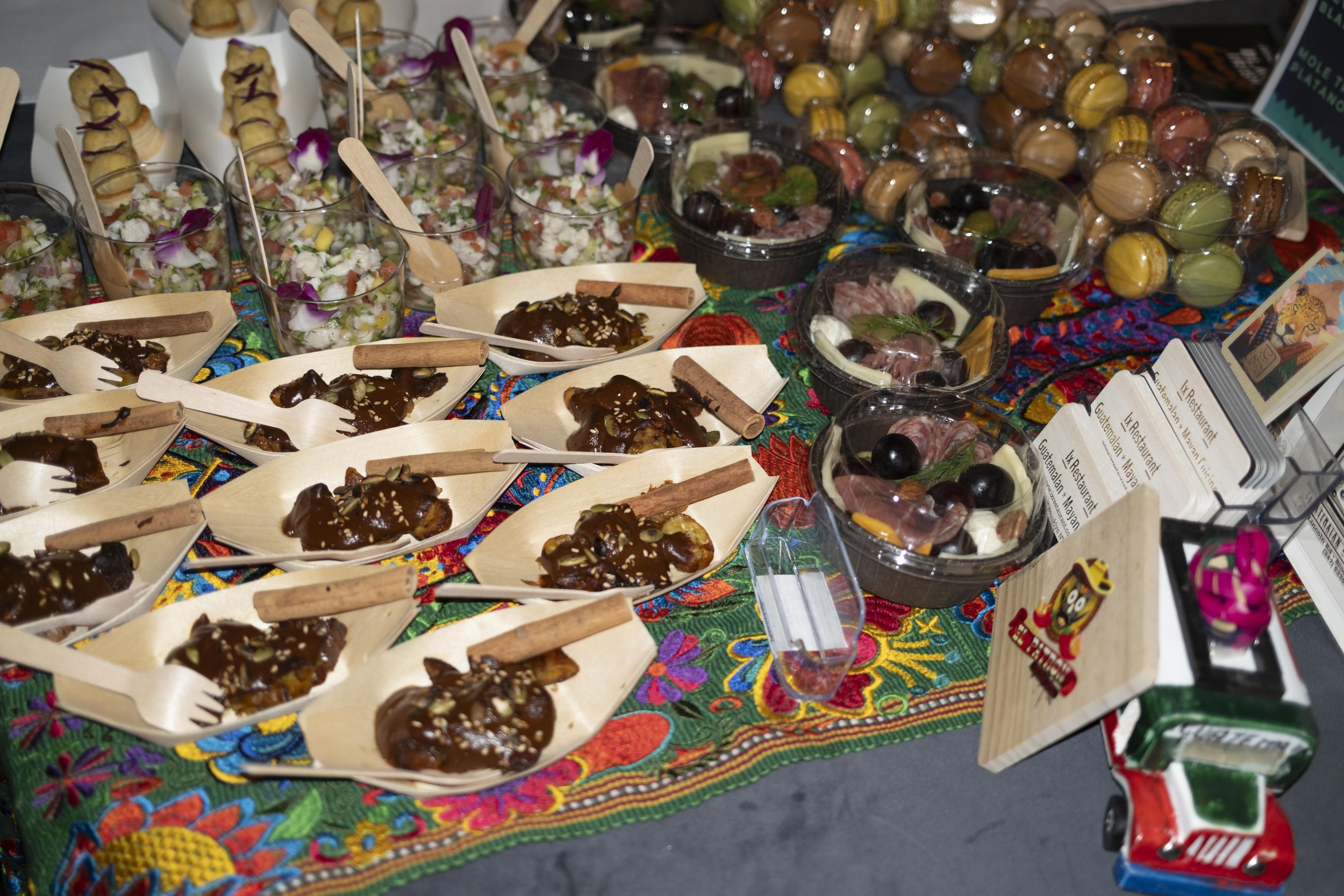 A colorful table displaying Mexican food and snacks, including mini tacos, fruit cups, and assorted candies, with a vibrant embroidered tablecloth.