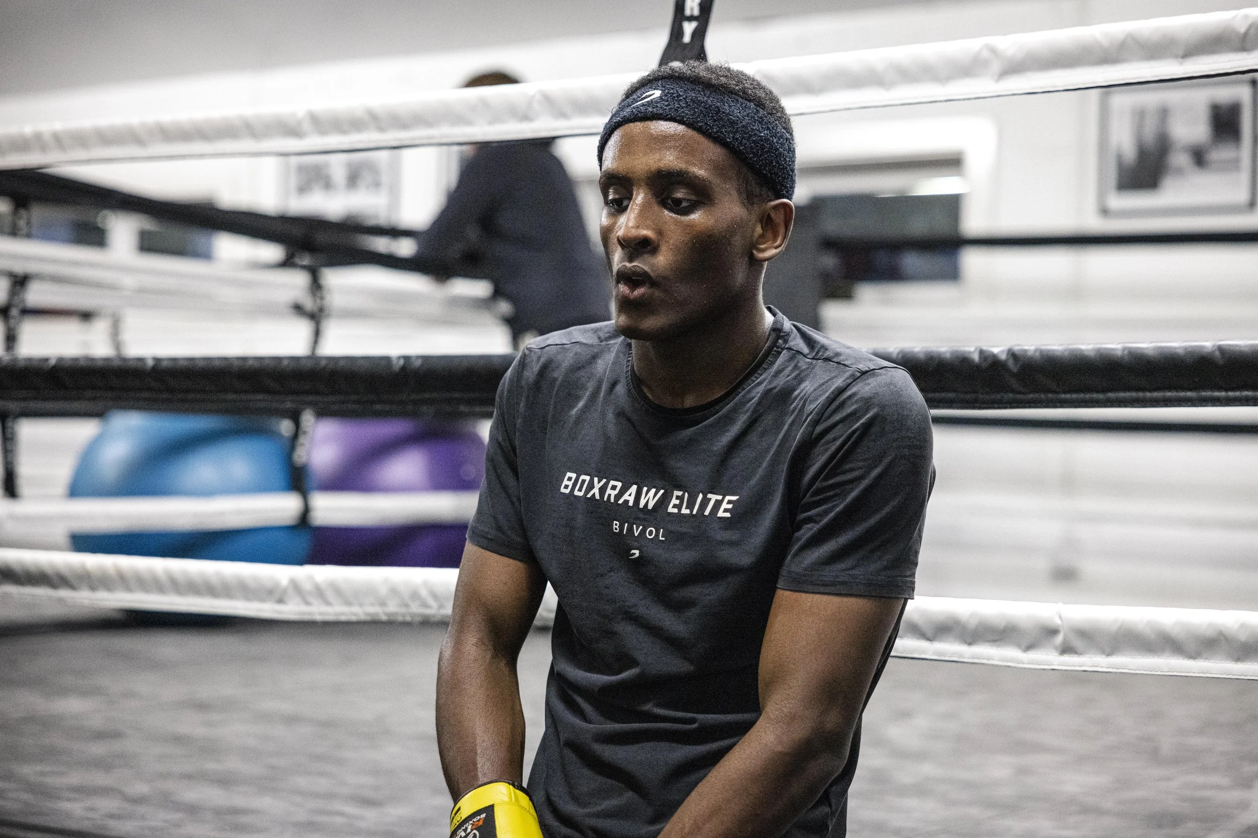 A young man in a black t-shirt and headband sits inside a boxing gym, with a boxing ring and exercise balls in the background.