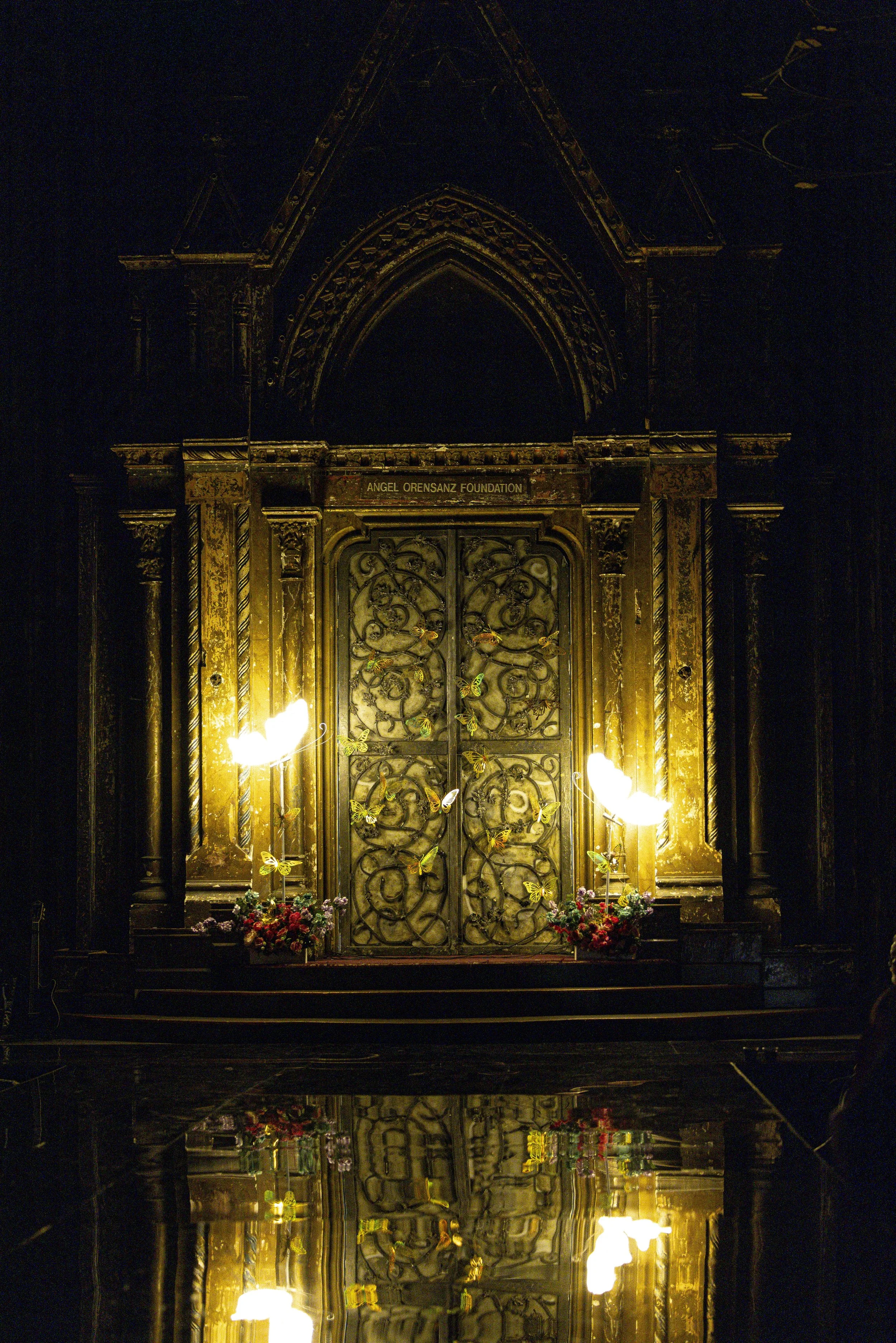 An ornate church altar with a gothic arch and intricate metalwork doors, illuminated by warm yellow lighting, with floral arrangements at the base and a sign reading 'Angel Orensanz Foundation' above the doors.