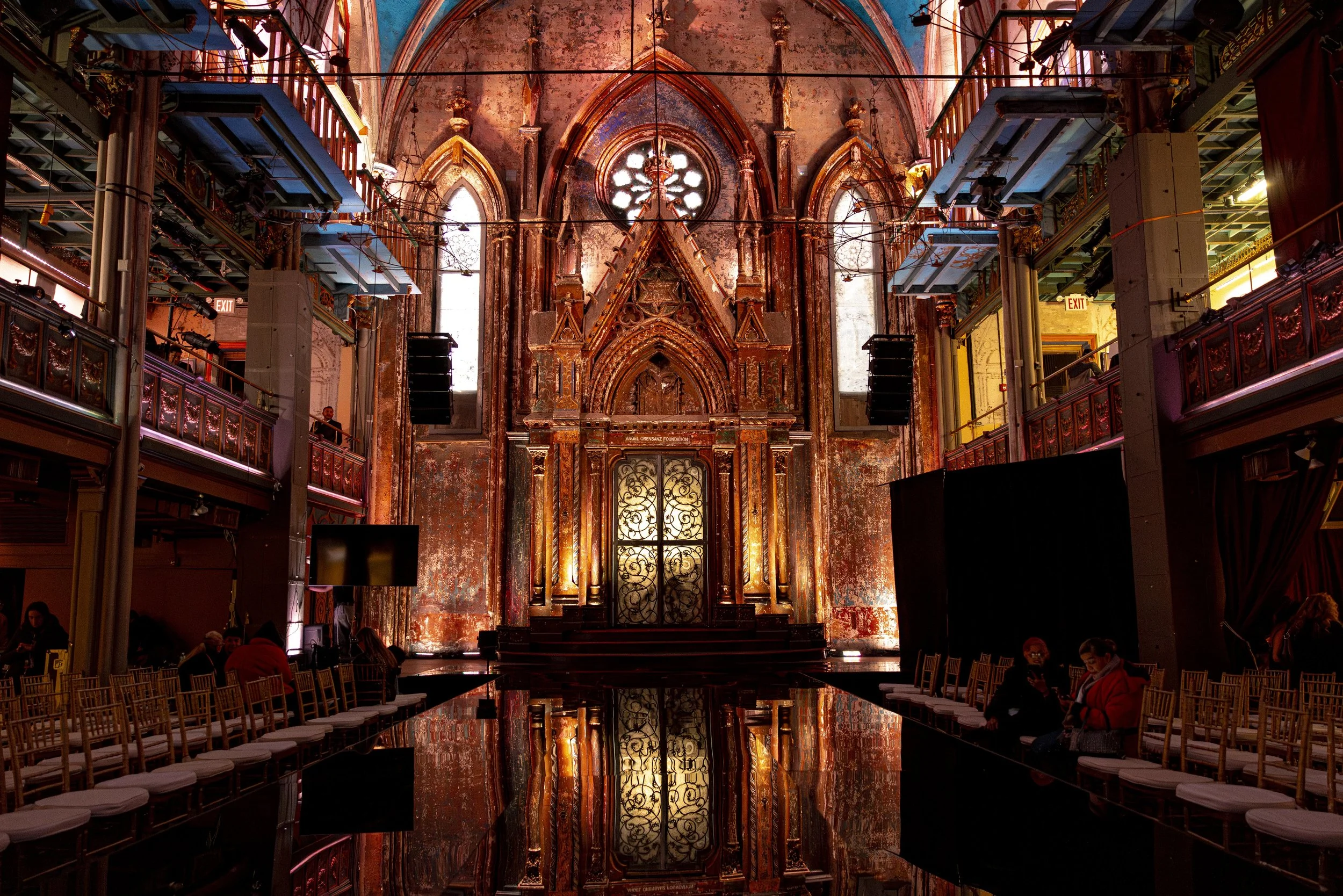 Interior of an old, abandoned church with Gothic architecture, high arched windows, and a large ornate altar. The walls are weathered and peeling, with some modern scaffolding and lighting equipment set up in the space. There are chairs arranged alon