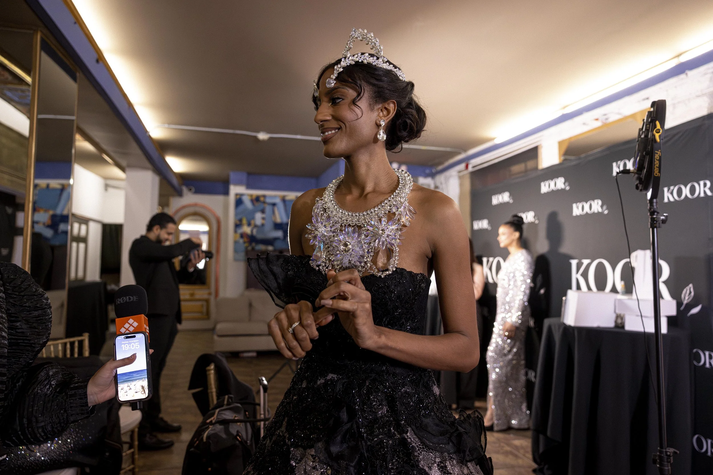 A woman dressed as a beauty queen, wearing a diamond tiara, earrings, and necklace, standing at a promotional event with a black background and the word 'KOOR' on it.